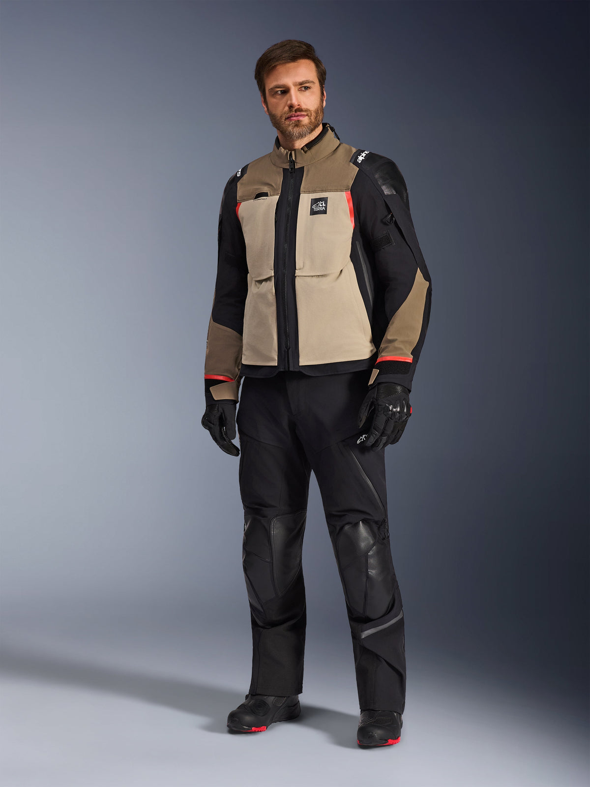 Un modelo masculino vistiendo la chaqueta Alpinestars Andes Pro Drystar® XF en Walnut Aluminum, combinada con pantalones, guantes y botas de motociclismo negros, con un diseño de múltiples paneles en color tostado y negro con acentos rojos para conducción de aventura.