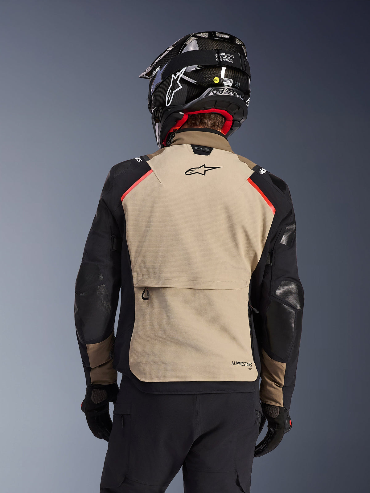 Chaqueta Alpinestars Andes Pro Drystar® XF en Walnut Aluminum, vista posterior, panel trasero de color canela con un gran bolsillo tipo cargo, mangas negras con paneles reforzados, logotipo de Alpinestars en la parte superior de la espalda, combinada con casco y guantes negros para touring de aventura.