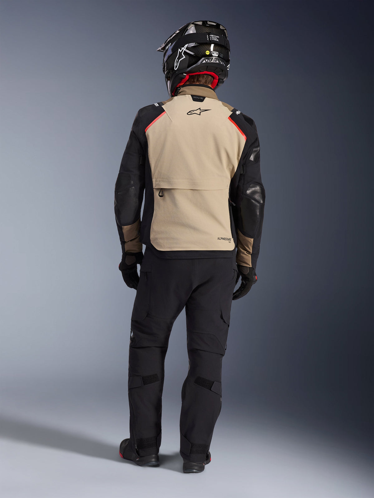 Motociclista de espaldas con la chaqueta Alpinestars Andes Pro Drystar® XF en Walnut Aluminum, panel trasero beige con mangas negras y detalles rojos, combinada con pantalones técnicos de aventura negros y casco de moto, equipamiento de touring premium para cualquier clima.