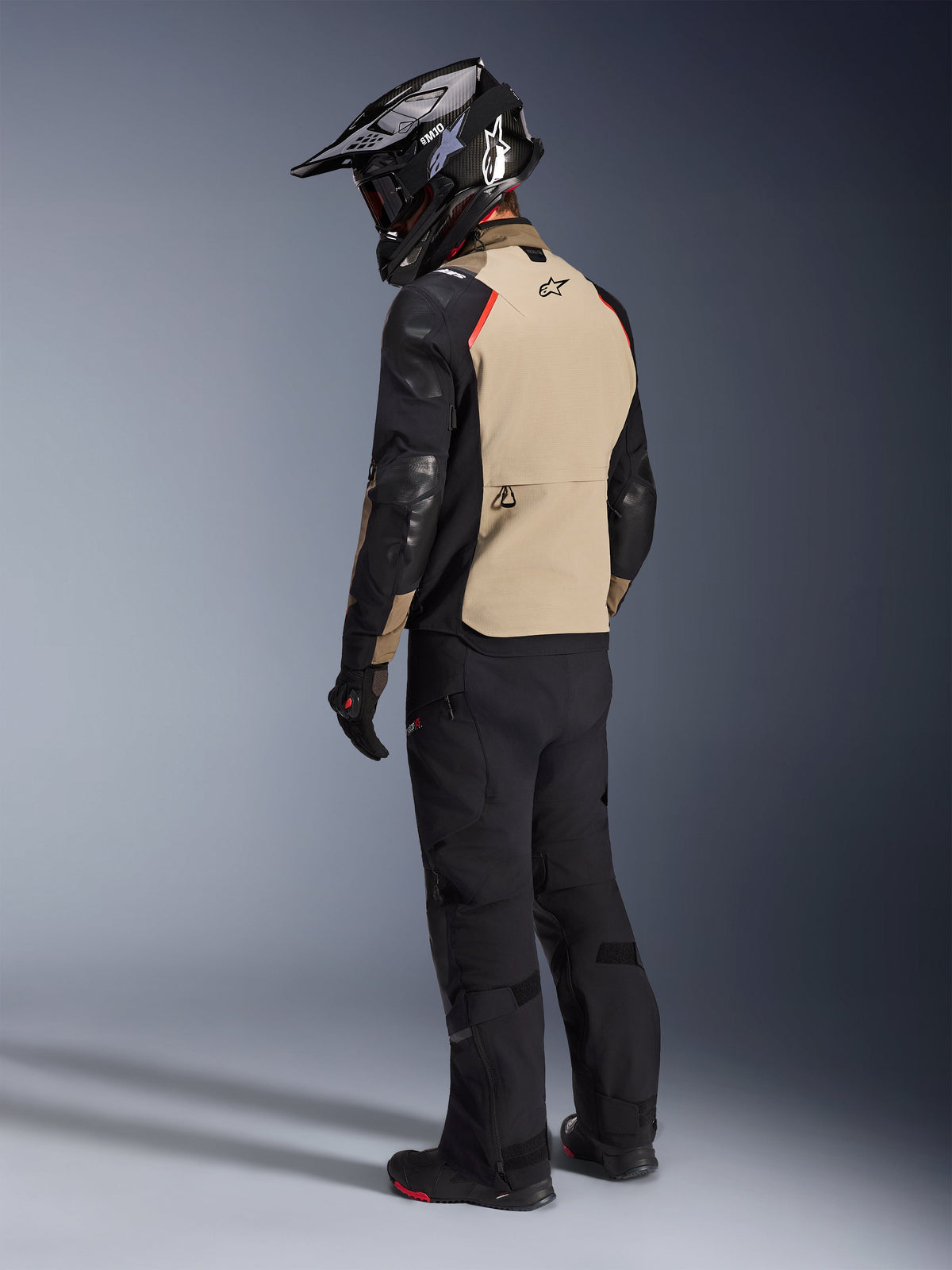 Motociclista visto desde atrás vistiendo la chaqueta Alpinestars Andes Pro Drystar® XF en Walnut Aluminum, con un panel trasero color canela, mangas negras y detalles rojos, casco de aventura negro, pantalones de motociclismo negros y botas negras, equipamiento profesional de adventure touring.