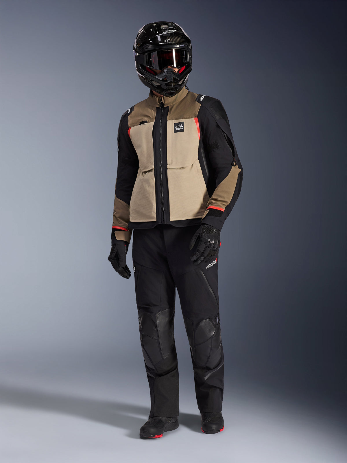 Chaqueta Alpinestars Andes Pro Drystar® XF en Walnut Aluminum, chaqueta de motociclismo de aventura combinada con pantalones negros a juego, casco adventure negro con gafas y botas negras, con paneles en color arena y negro con detalles en rojo.