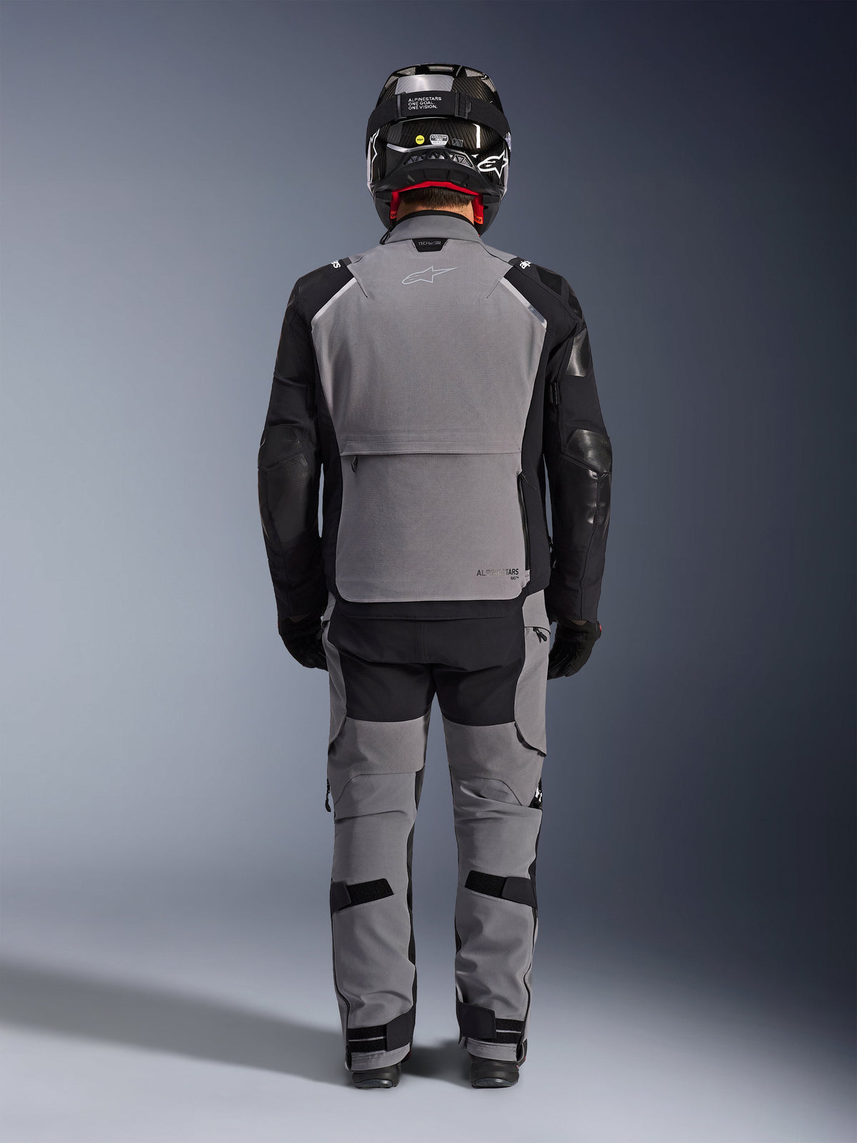 Chaqueta Alpinestars Andes Pro Drystar® XF, vista trasera, equipamiento de aventura touring en gris oscuro y negro, usada con pantalones grises y negros a juego, casco de fibra de carbono negro y guantes negros, con paneles técnicos y logotipos reflectantes