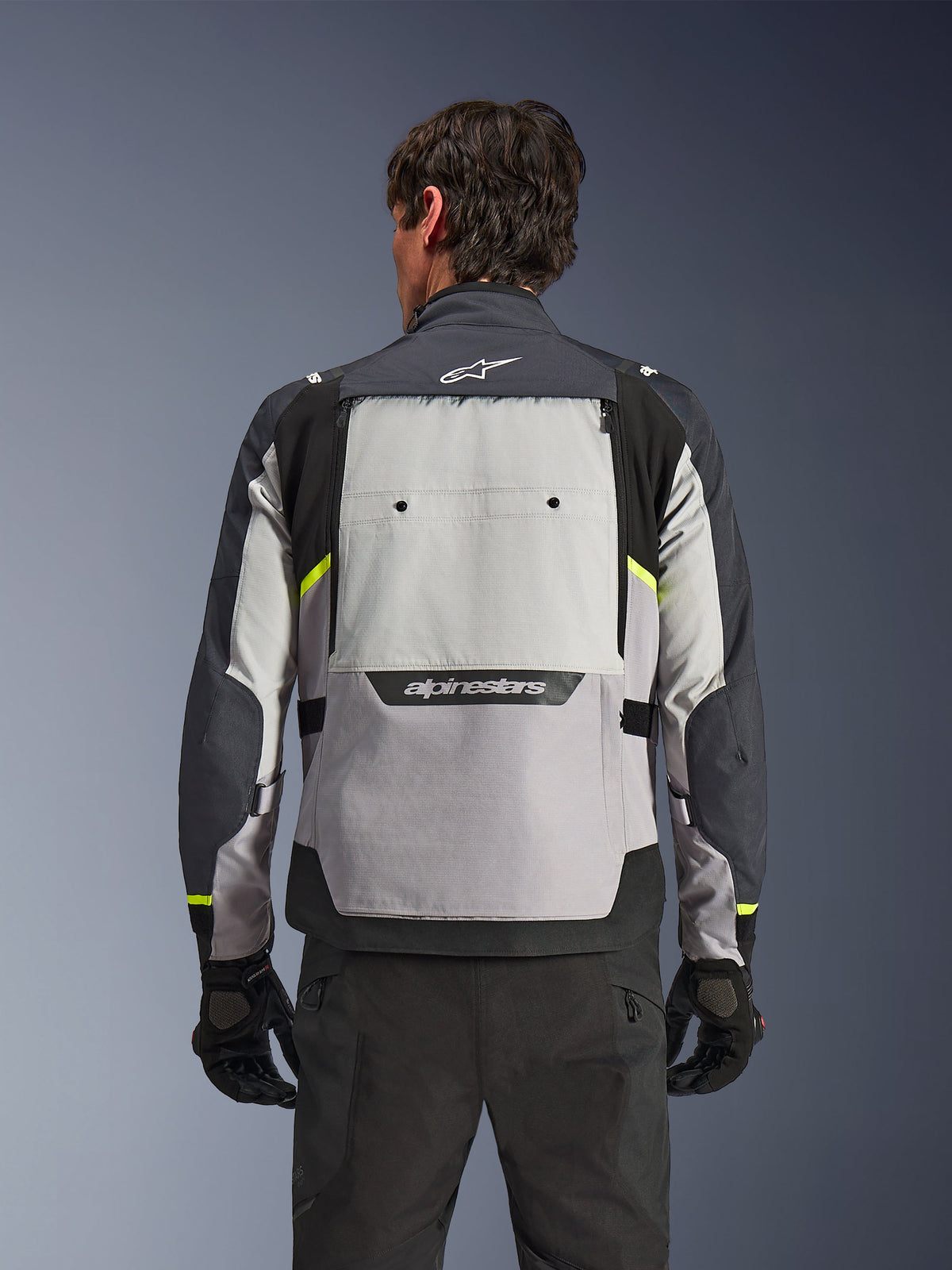 Vista trasera de un motociclista con la chaqueta Alpinestars Cusco Drystar® en colores Ice Gray, Mid Gray y Deep Gray, con detalles en amarillo neón, un panel de ventilación grande en la espalda y logotipos destacados de Alpinestars, combinada con pantalones de motociclista y guantes negros.