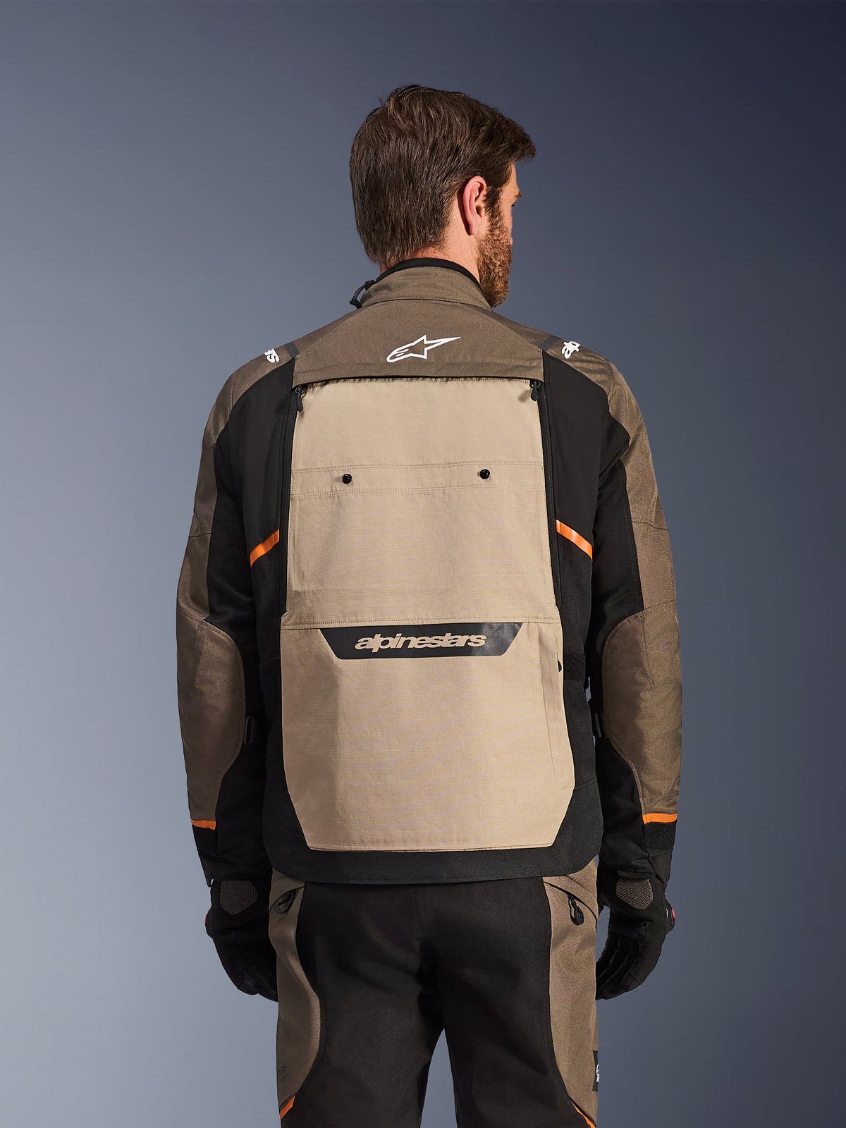 Chaqueta Alpinestars Cusco Drystar®, Walnut Aluminum Black, vista trasera, con un gran panel posterior color canela con logotipo negro, acentos naranjas, usada con pantalones y guantes a juego, diseñada para el turismo de aventura.