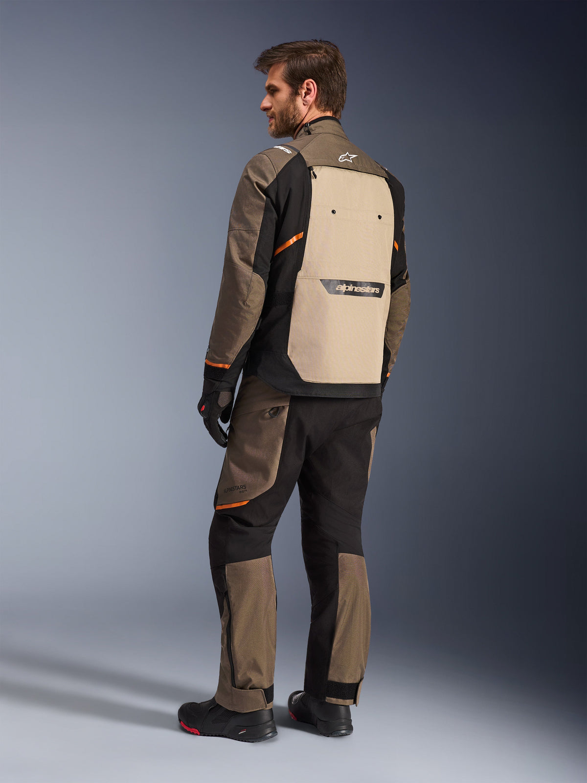 Chaqueta Alpinestars Cusco Drystar® en color Walnut Aluminum Black, vista trasera que muestra el panel posterior en color arena y detalles en naranja, usada con pantalones de moto a juego, guantes y botas negras, equipamiento profesional de adventure touring
