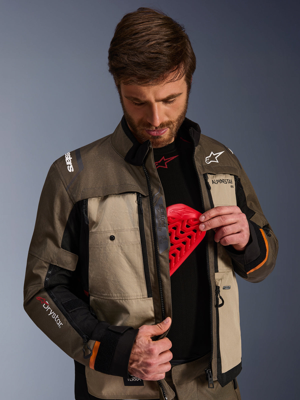Chaqueta Alpinestars Cusco Drystar®, Walnut Aluminum Black, usada por un hombre que muestra el bolsillo para el protector de pecho con una inserción de armadura roja, chaqueta textil de motociclismo con ventilaciones con cremallera y logotipos para adventure touring.