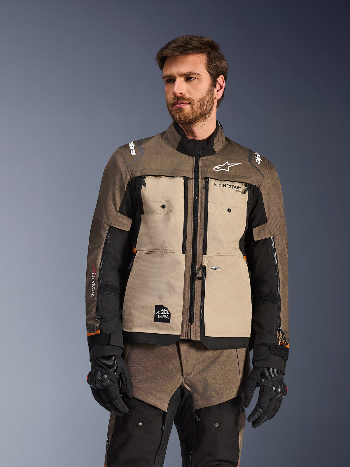 Chaqueta Alpinestars Cusco Drystar®, chaqueta de motociclismo adventure, nogal, aluminio y negro, usada por un modelo con guantes negros, con paneles frontales en color arena y mangas negras con logotipos de Alpinestars y parche All Terra, equipamiento de touring protector para todo clima