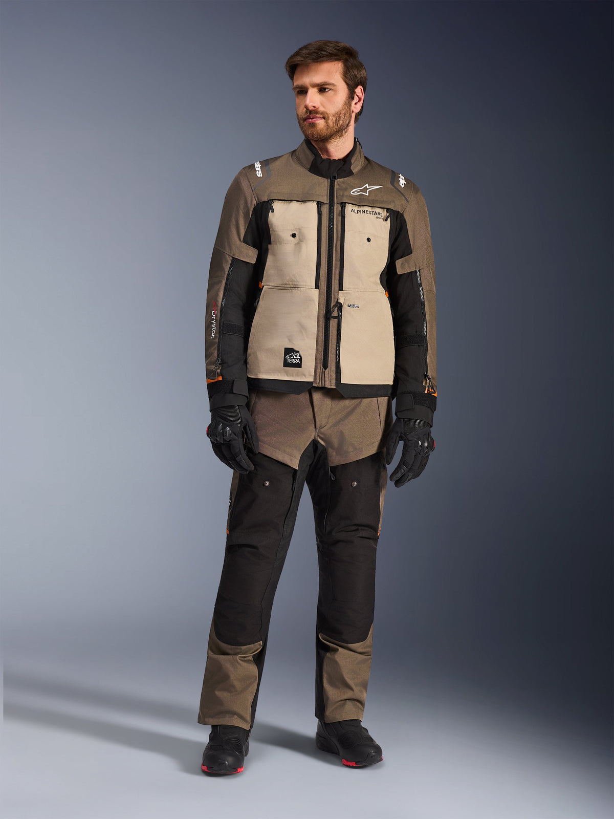 Chaqueta Alpinestars Cusco Drystar® en color Walnut Aluminum Black, vestida por un piloto masculino con pantalones de aventura touring a juego, guantes negros y botas, con paneles beige en el pecho, mangas negras con sutiles detalles en naranja y cremalleras de ventilación funcionales para la conducción en carretera.