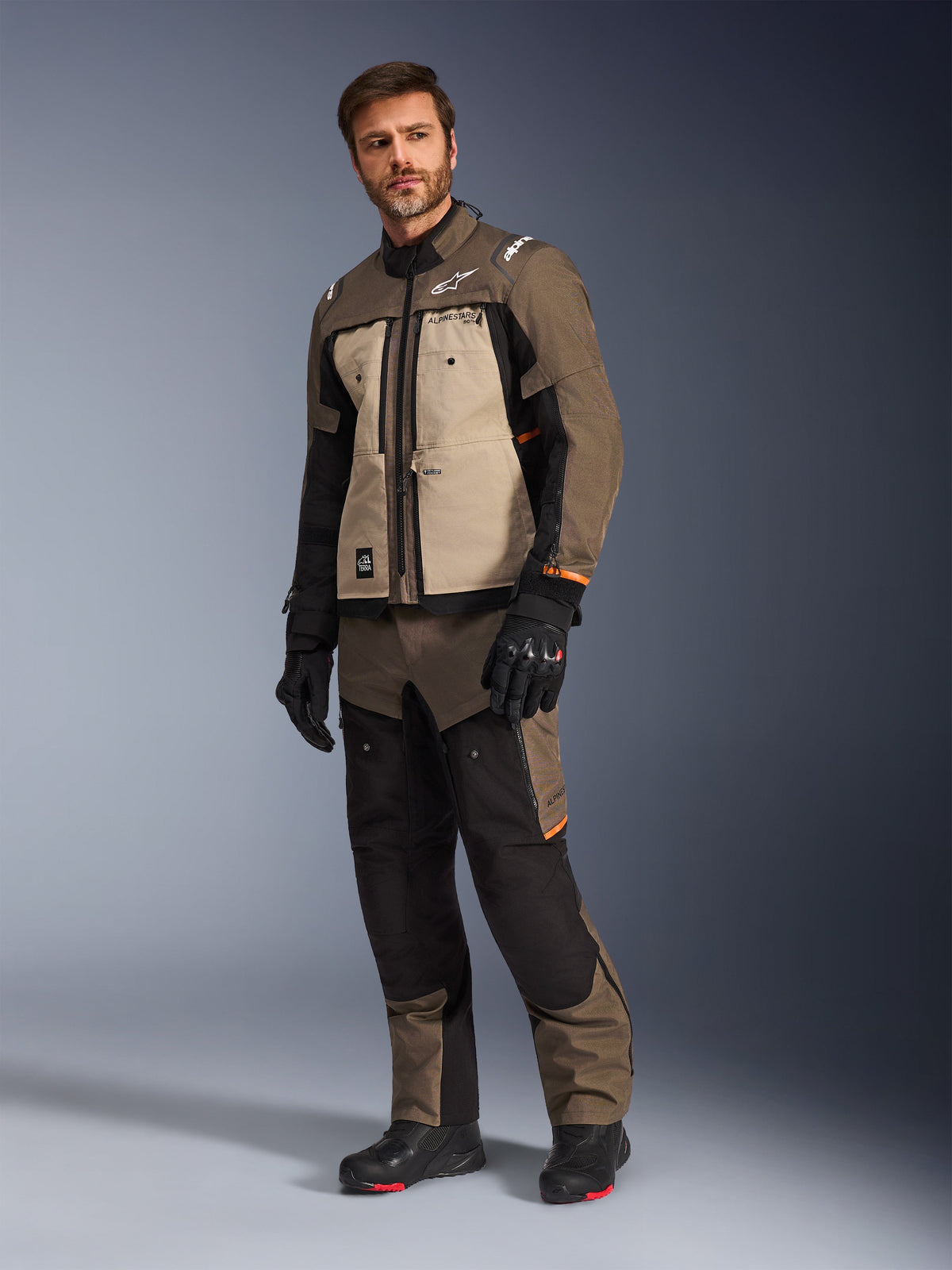 Chaqueta Alpinestars Cusco Drystar® en Walnut Aluminum Black, lucida por un modelo masculino con pantalones y guantes de moto a juego, diseño de turismo de aventura con paneles de color canela en el pecho y detalles en negro.