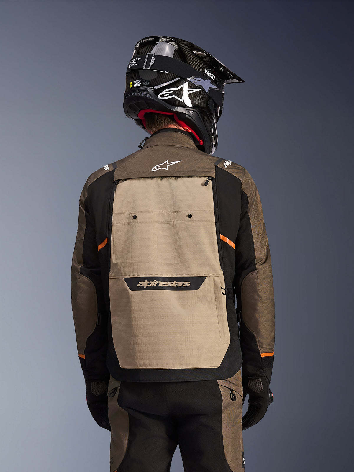 Vista trasera de la chaqueta Alpinestars Cusco Drystar® en color Walnut Aluminum Black, chaqueta de motociclismo adventure touring con un panel posterior grande en color tostado y el logotipo de Alpinestars, mangas negras con detalles en naranja, mostrada con un casco estilo carbono negro.
