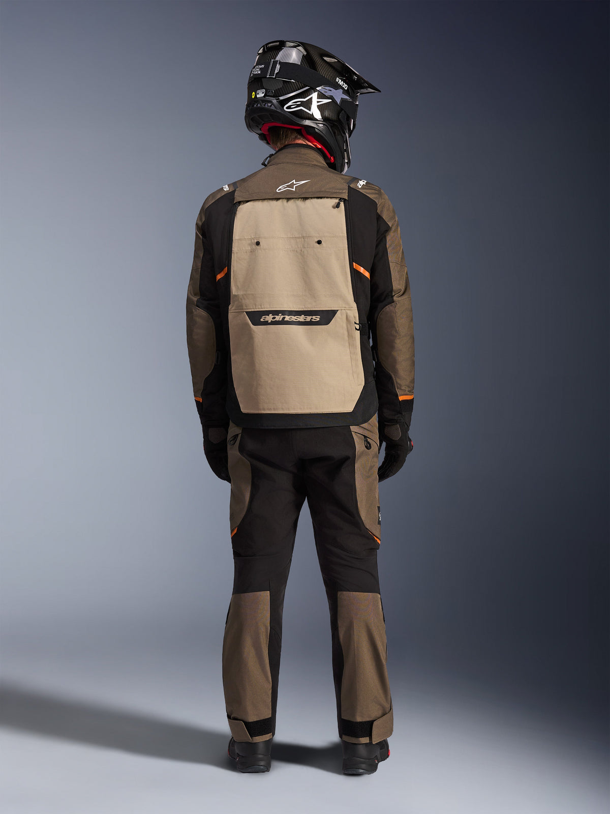 Motorista visto desde atrás con la chaqueta Alpinestars Cusco Drystar® en Walnut Aluminum Black, con un gran bolsillo trasero color aluminio y detalles en naranja, pantalones adventure a juego, casco off-road negro y guantes, equipamiento textil resistente para touring.