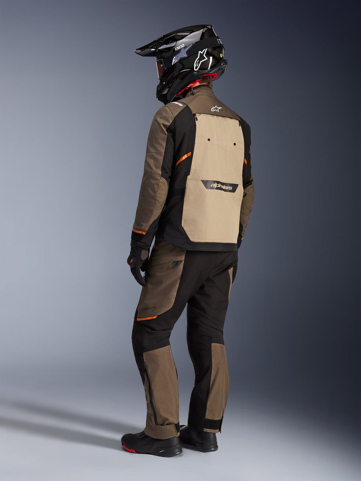 Chaqueta Alpinestars Cusco Drystar®, chaqueta de turismo de aventura, Walnut Aluminum Black, vista posterior que muestra el panel trasero en color canela y detalles en naranja, combinada con pantalones a juego y casco de fibra de carbono negro.