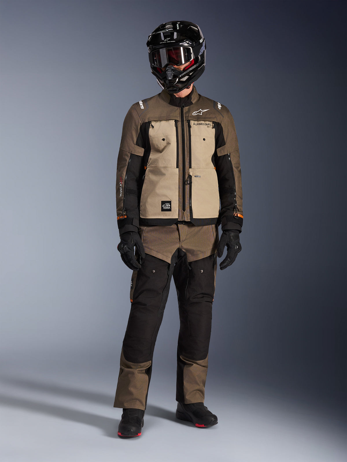 Chaqueta Alpinestars Cusco Drystar®, Walnut Aluminum Black, chaqueta de motociclismo adventure touring usada con pantalones a juego y casco negro, con paneles frontales color canela y mangas de ripstop marrón, equipamiento técnico para todo tipo de clima
