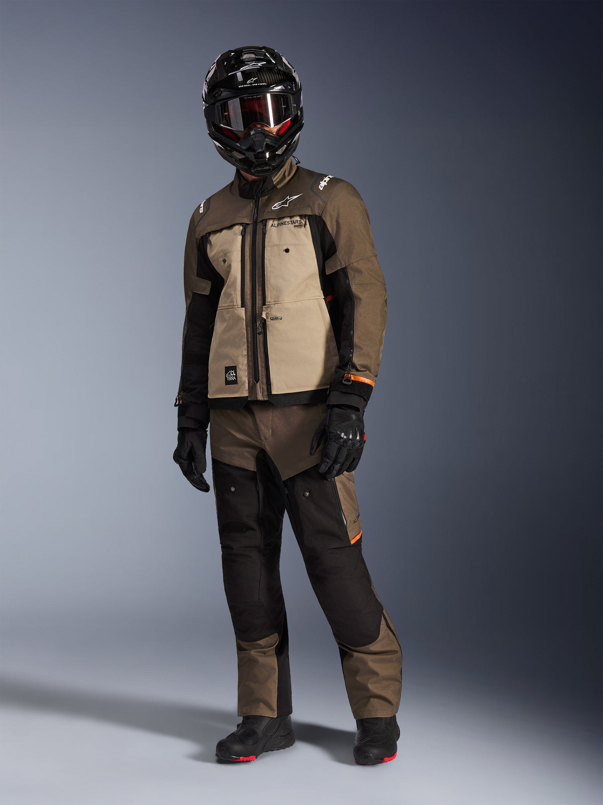 Chaqueta Alpinestars Cusco Drystar®, Walnut Aluminum Black, vestida por un piloto con equipamiento completo de adventure touring, con paneles en tonos canela y marrón con detalles negros, coordinada con pantalones a juego, casco integral negro con gafas y botas negras, fondo de estudio.