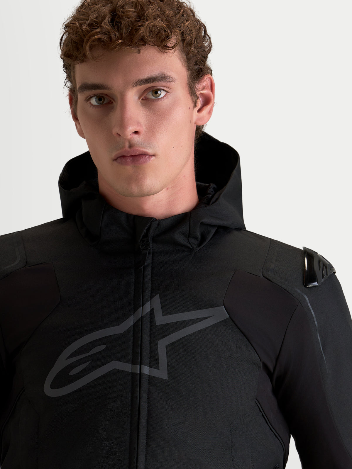Alpinestars Zaca V 2 Waterproof, chaqueta de moto negra impermeable con capucha integrada y protecciones DFS en los hombros, retrato de primer plano en modelo