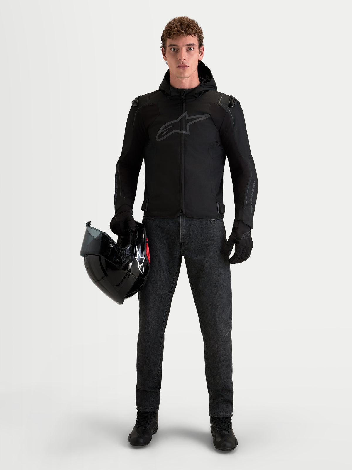 Alpinestars Zaca V 2 Waterproof Jacket, chaqueta de moto impermeable negra con capucha para hombre, vestida por un piloto de pie sosteniendo un casco