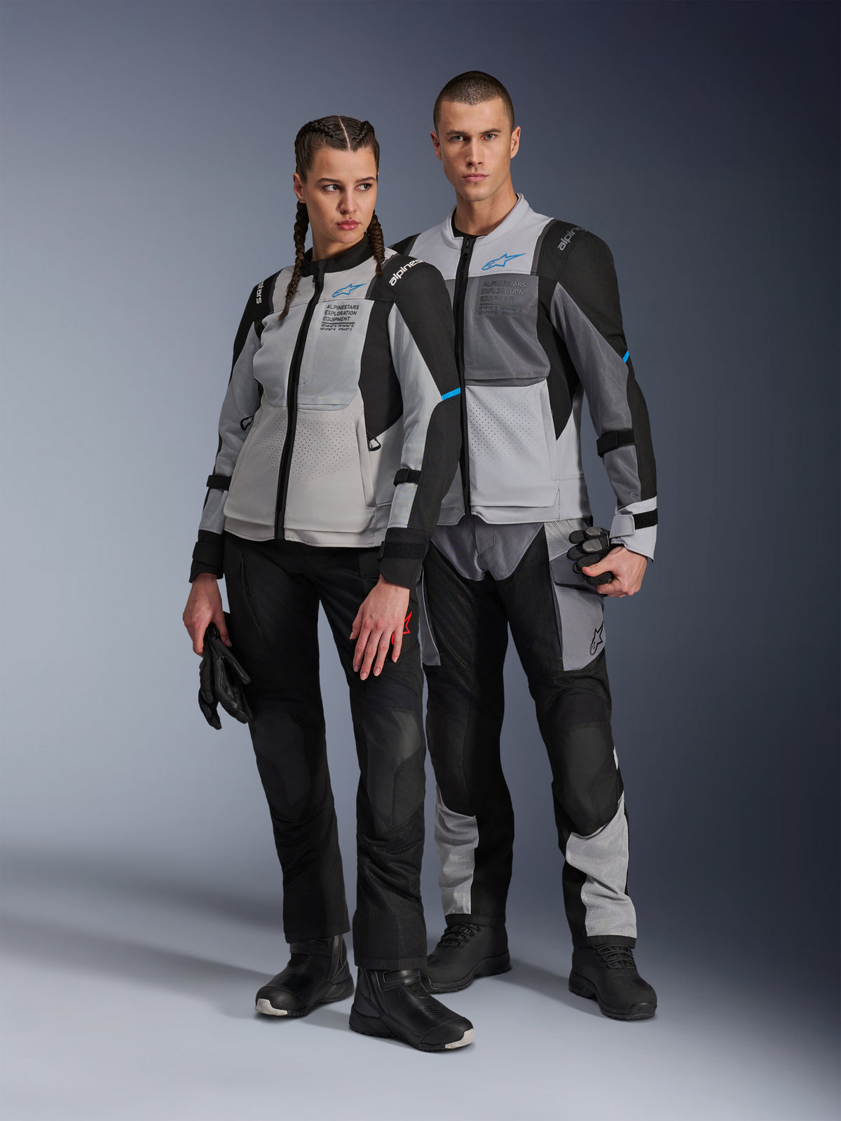 Man and woman wearing Alpinestars Stella ST-2 Air Jackets and matching technical pants, Ice Gray and Black colorway with blue accents, featuring mesh ventilation and 'ALPINESTARS EXPLORATION EQUIPMENT' branding, studio couple shot