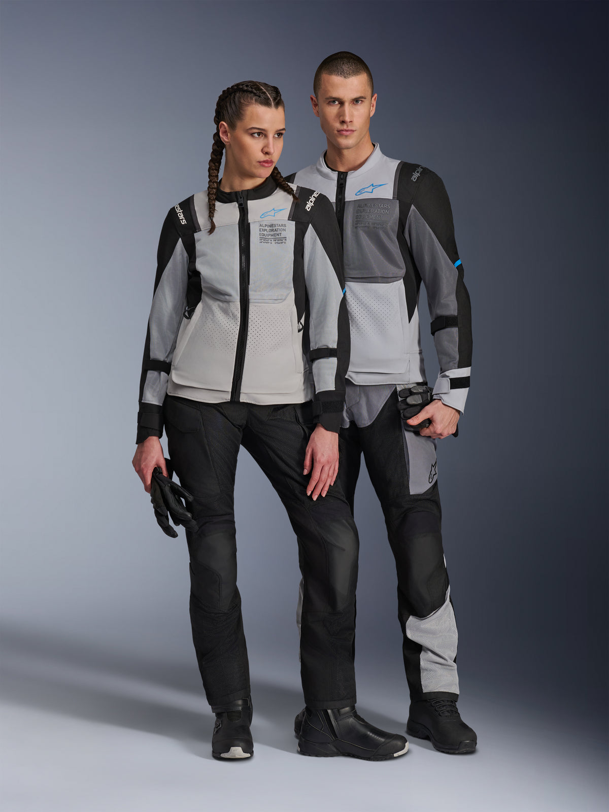 Male and female models wearing Alpinestars ST-2 Air Jackets in Ice Gray Dark Gray and Black, technical summer touring motorcycle gear featuring large mesh panels and exploration equipment branding, paired with black riding pants, gloves, and boots.