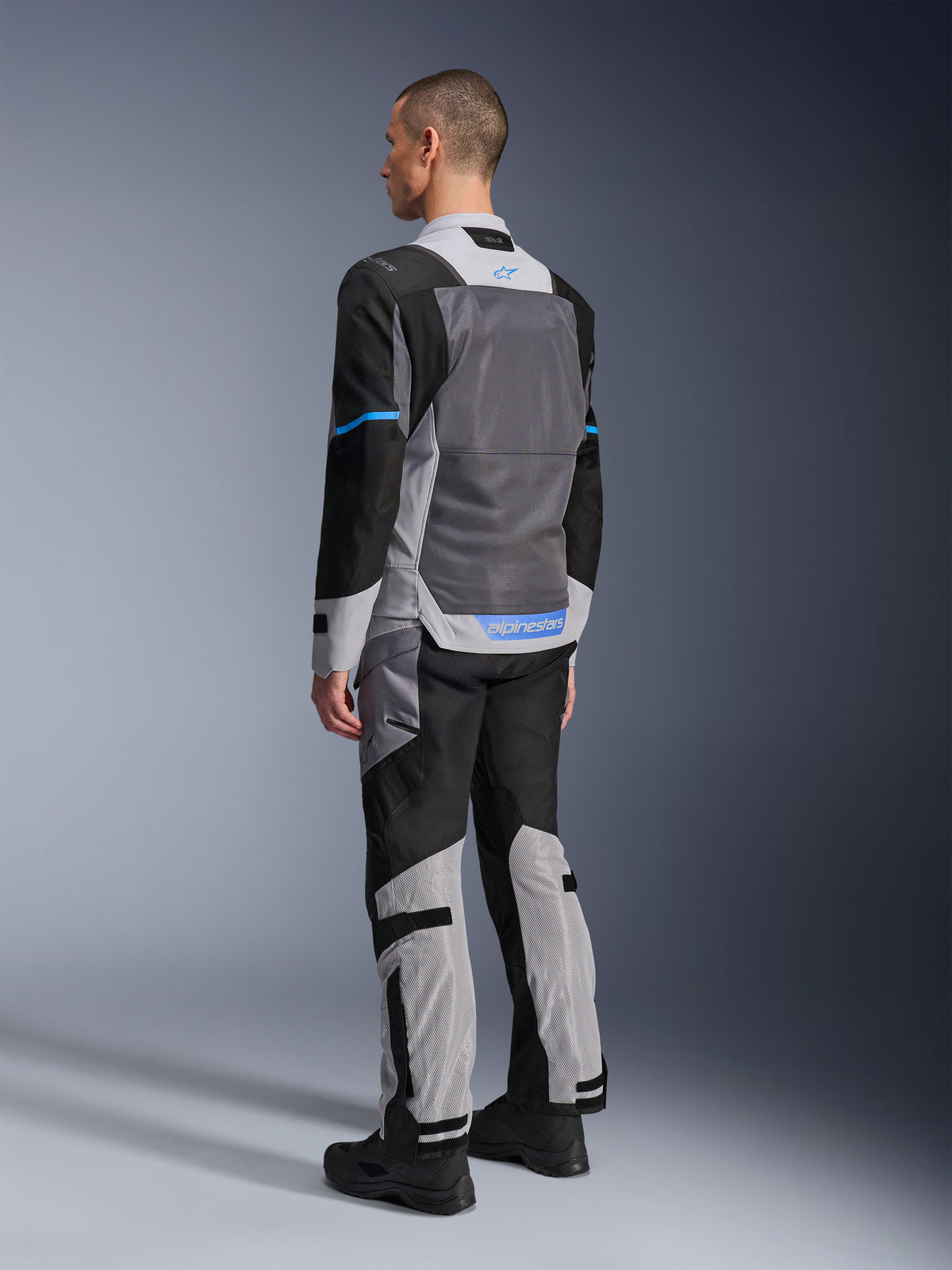 Modelo masculino de espaldas luciendo la chaqueta Alpinestars ST-2 Air en gris hielo, gris oscuro y negro, con un gran panel de malla gris en la espalda, detalles del logo en azul y pantalones de moto a juego para turismo de verano.