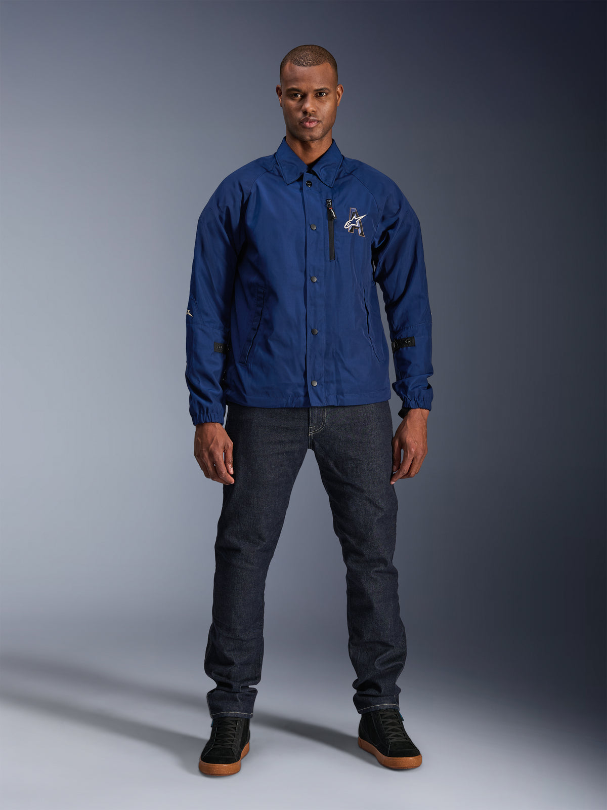 Chaqueta Alpinestars Revved WR, azul marino, chaqueta de moto de estilo coach, vestida por un hombre con vaqueros oscuros y zapatillas negras, con cierre de botones a presión y logotipo de Alpinestars en 3D, estilo urbano contemporáneo