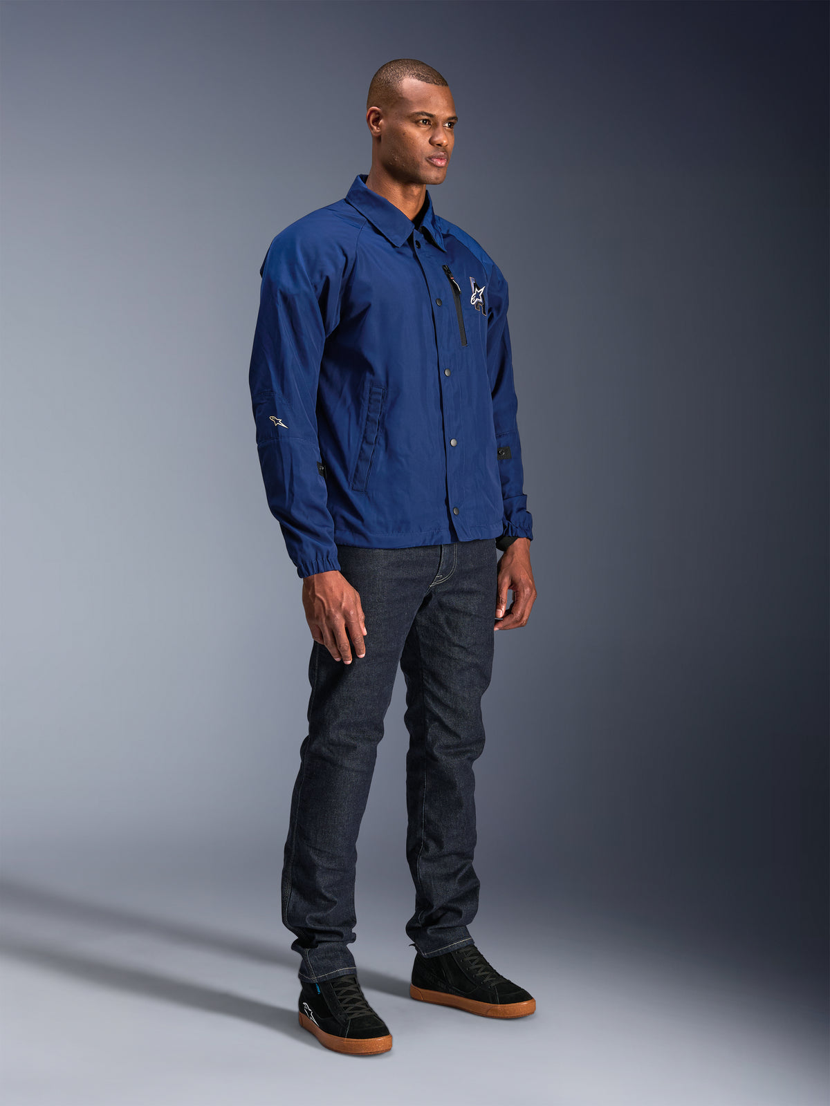 Chaqueta Alpinestars Revved WR, chaqueta de moto azul, lucida por un modelo masculino con vaqueros de denim oscuro y zapatillas negras, con silueta de chaqueta tipo coach resistente al agua, botones a presión frontales y logotipo minimalista en el pecho.