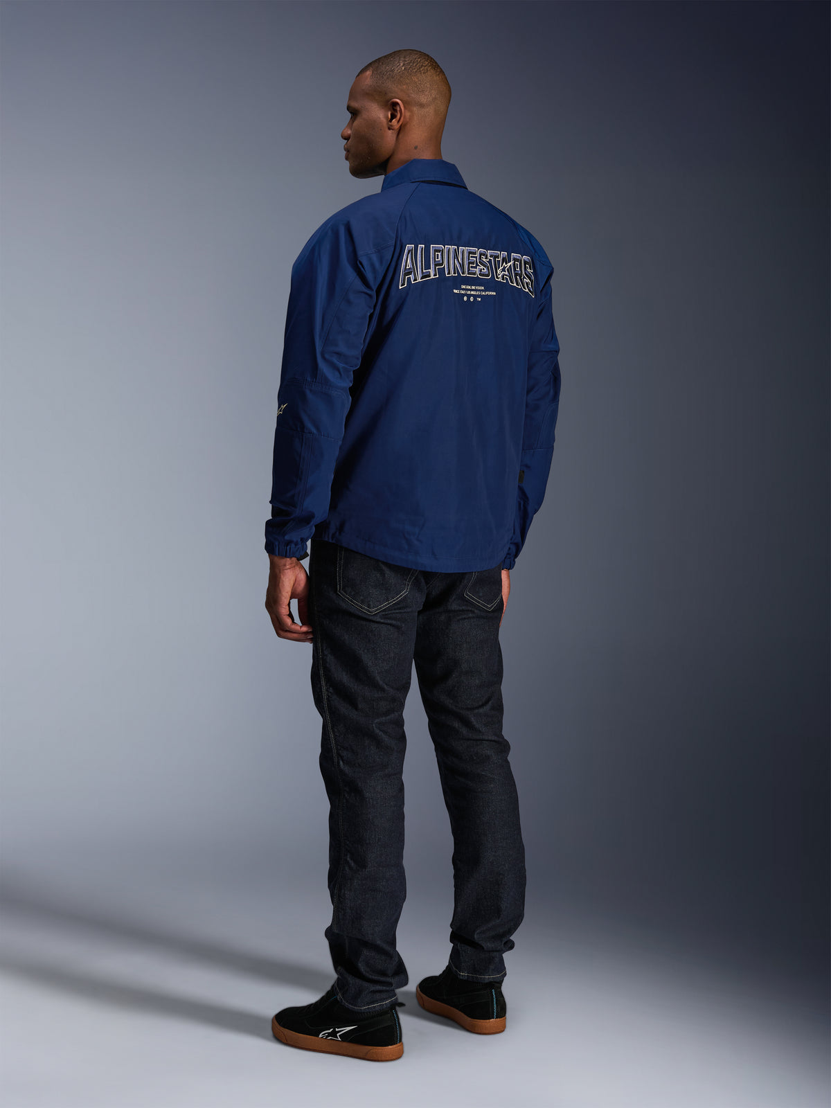 Modelo luciendo la chaqueta Alpinestars Revved WR en azul marino desde atrás, con un gran bordado de 'ALPINESTARS' perfilado en blanco en la espalda, combinada con pantalones vaqueros oscuros y zapatillas negras, estética de motociclismo urbano.