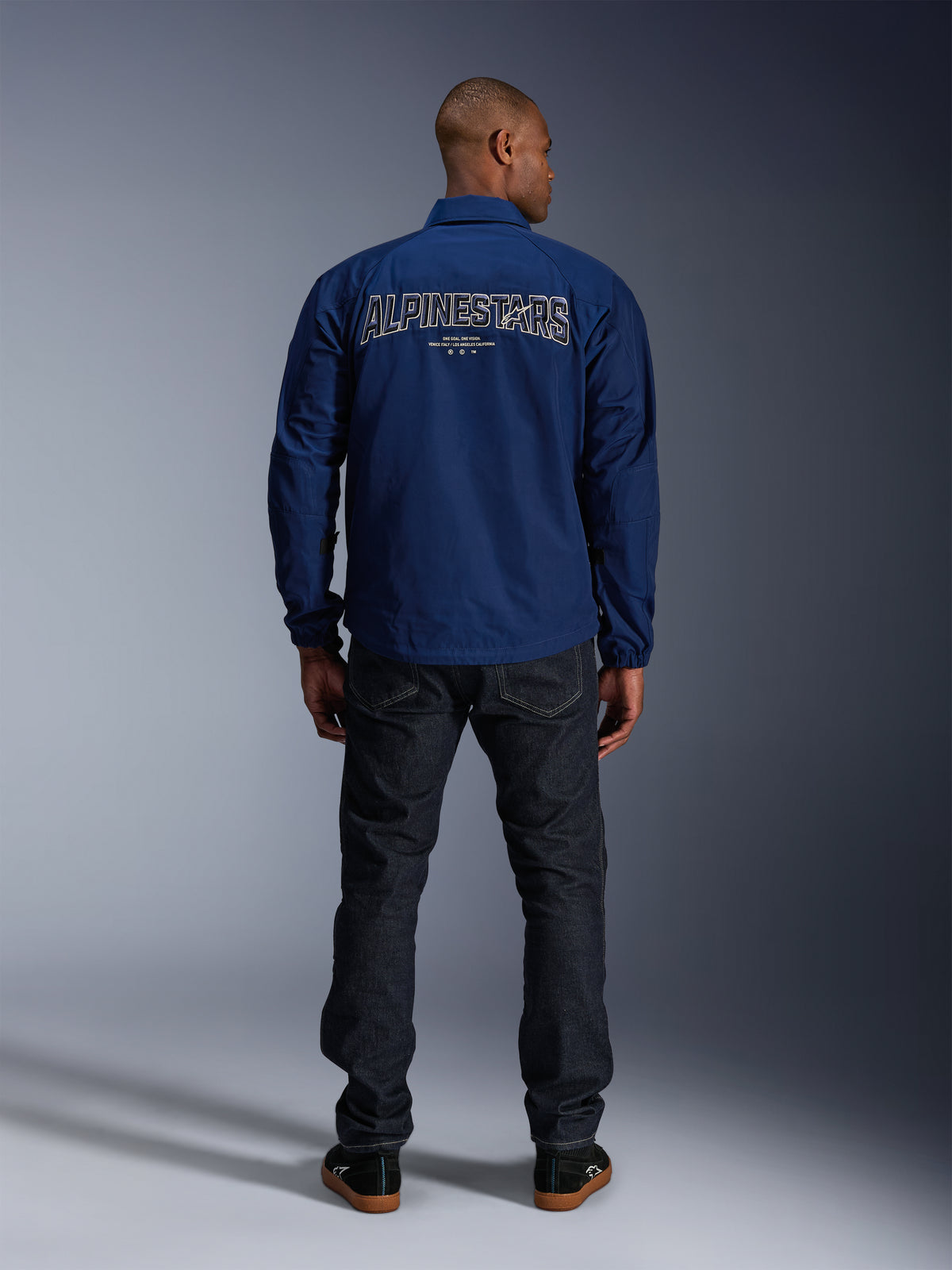 Chaqueta Alpinestars Revved WR en azul marino, vista trasera, con un gran bordado 'ALPINESTARS' con efecto 3D en la espalda, combinada con vaqueros de denim oscuro y zapatillas negras, ropa urbana para motociclista.