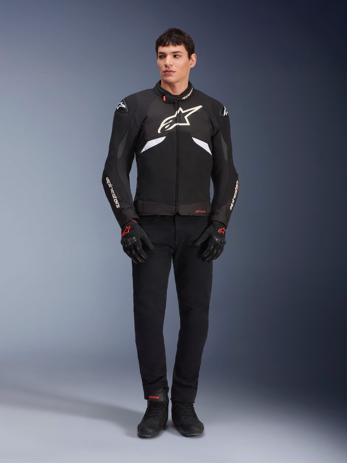 Modelo masculino luciendo la chaqueta Alpinestars T-GP R V3 Drystar® en negro y blanco, una chaqueta deportiva textil e impermeable con un gran logotipo blanco de Alpinestars en el pecho, combinada con pantalones y guantes negros; retrato de estudio profesional que resalta su estilizada silueta deportiva.