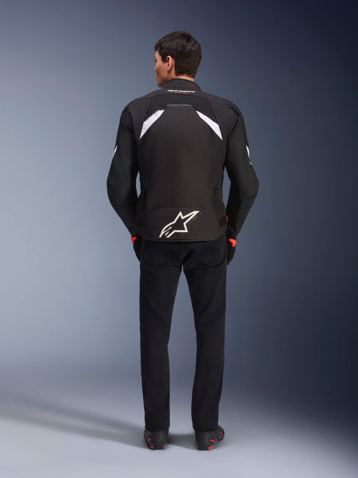 Chaqueta Alpinestars T-GP R V3 Drystar®, Negro y Blanco, vista posterior, chaqueta textil de moto impermeable con un gran logotipo de estrella blanca en la parte inferior de la espalda y detalles blancos en los hombros, combinada con pantalones y botas negros, diseño deportivo derivado de la competición