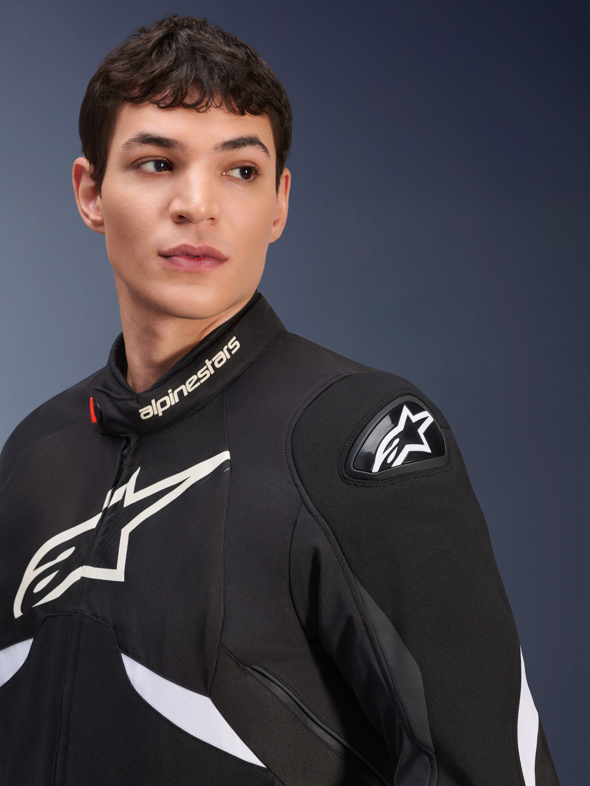 Chaqueta Alpinestars T-GP R V3 Drystar®, en negro y blanco, usada por un modelo masculino, con logotipos Alpinestars blancos en el cuello y el pecho, deslizador de hombro DFS negro con una estrella blanca, toma detallada de una chaqueta textil deportiva impermeable.