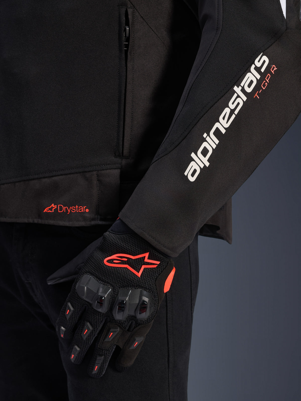 Chaqueta Alpinestars T-GP R V3 Drystar®, chaqueta de moto textil negra, vista de cerca que muestra los logotipos blancos y naranjas en la manga, logotipo Drystar naranja en el bajo, combinada con guantes de malla protectores negros con detalles rojos.