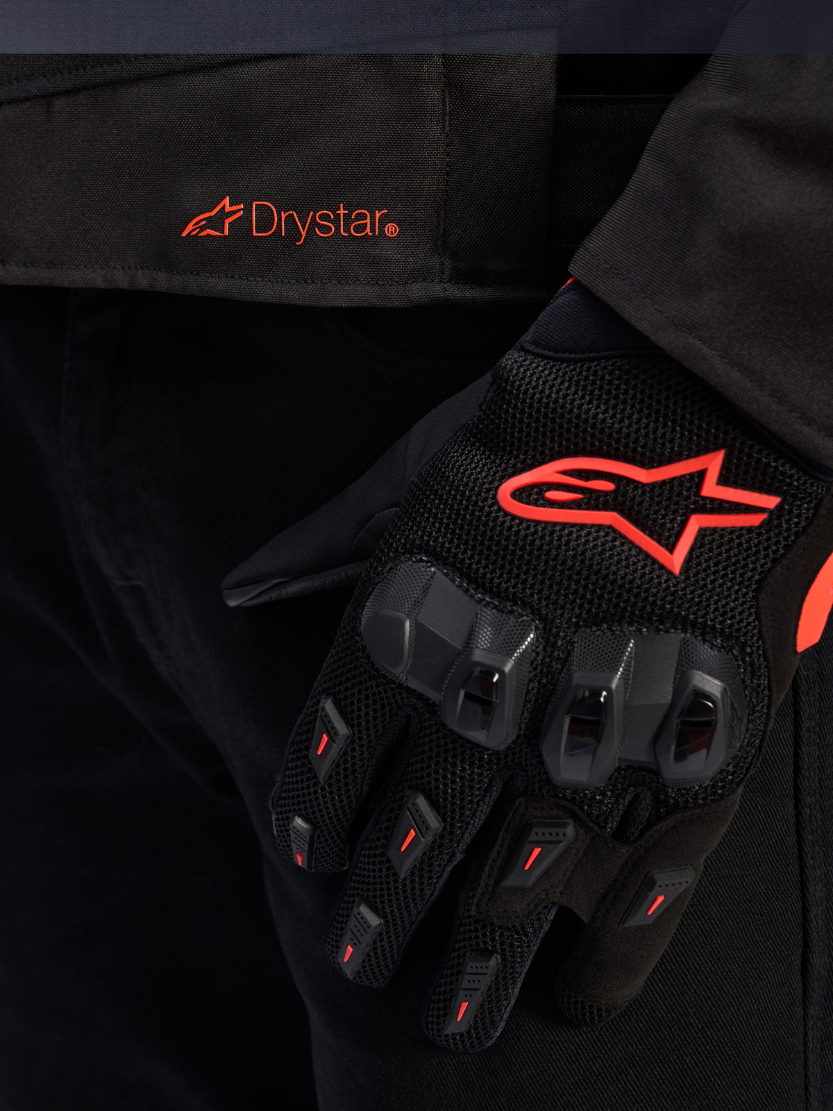 Primer plano de la cintura de la chaqueta Alpinestars T-GP R V3 Drystar® con el logotipo Drystar en rojo, mostrada con un guante de moto negro con el logotipo de la estrella de Alpinestars en rojo, tejido de malla y protección de nudillos reforzada.