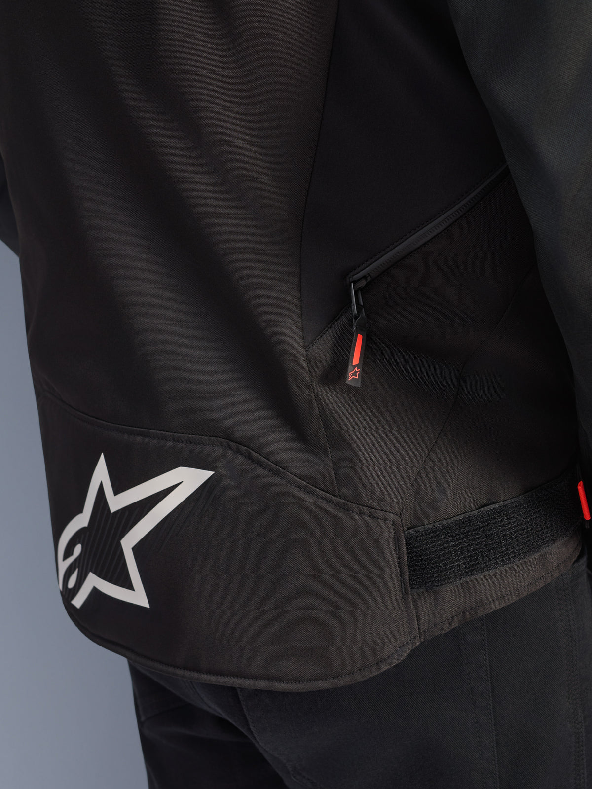 Chaqueta Alpinestars T-GP R V3 Drystar®, negro y blanco, primer plano de la parte inferior de la espalda con un gran logotipo de la estrella de Alpinestars en blanco, un bolsillo lateral con cremallera y tirador rojo, y una correa de ajuste de velcro en la cintura, material textil duradero.