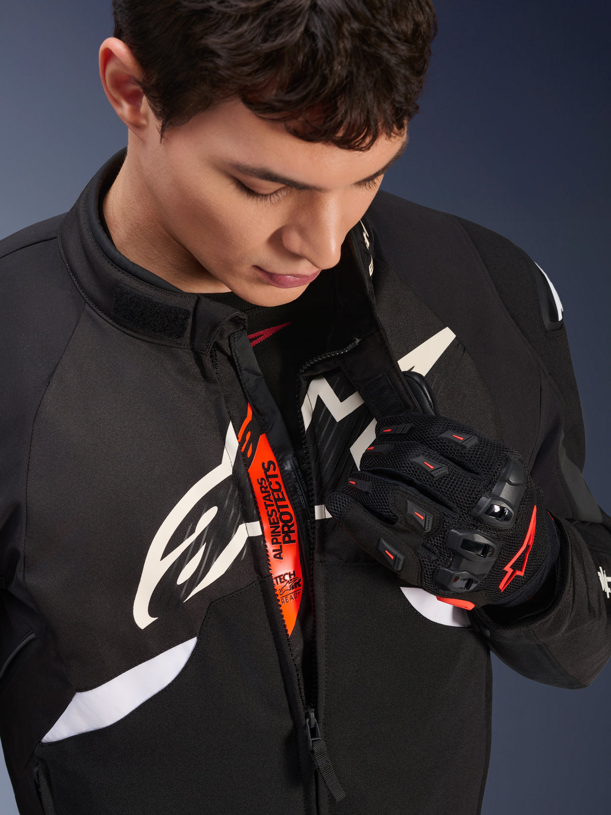 Chaqueta Alpinestars T-GP R V3 Drystar®, chaqueta textil deportiva para moto en blanco y negro, parcialmente abierta revelando la cinta interna naranja 'Alpinestars Protects', usada con guantes de malla negros y rojos, equipamiento de protección derivado de la competición.