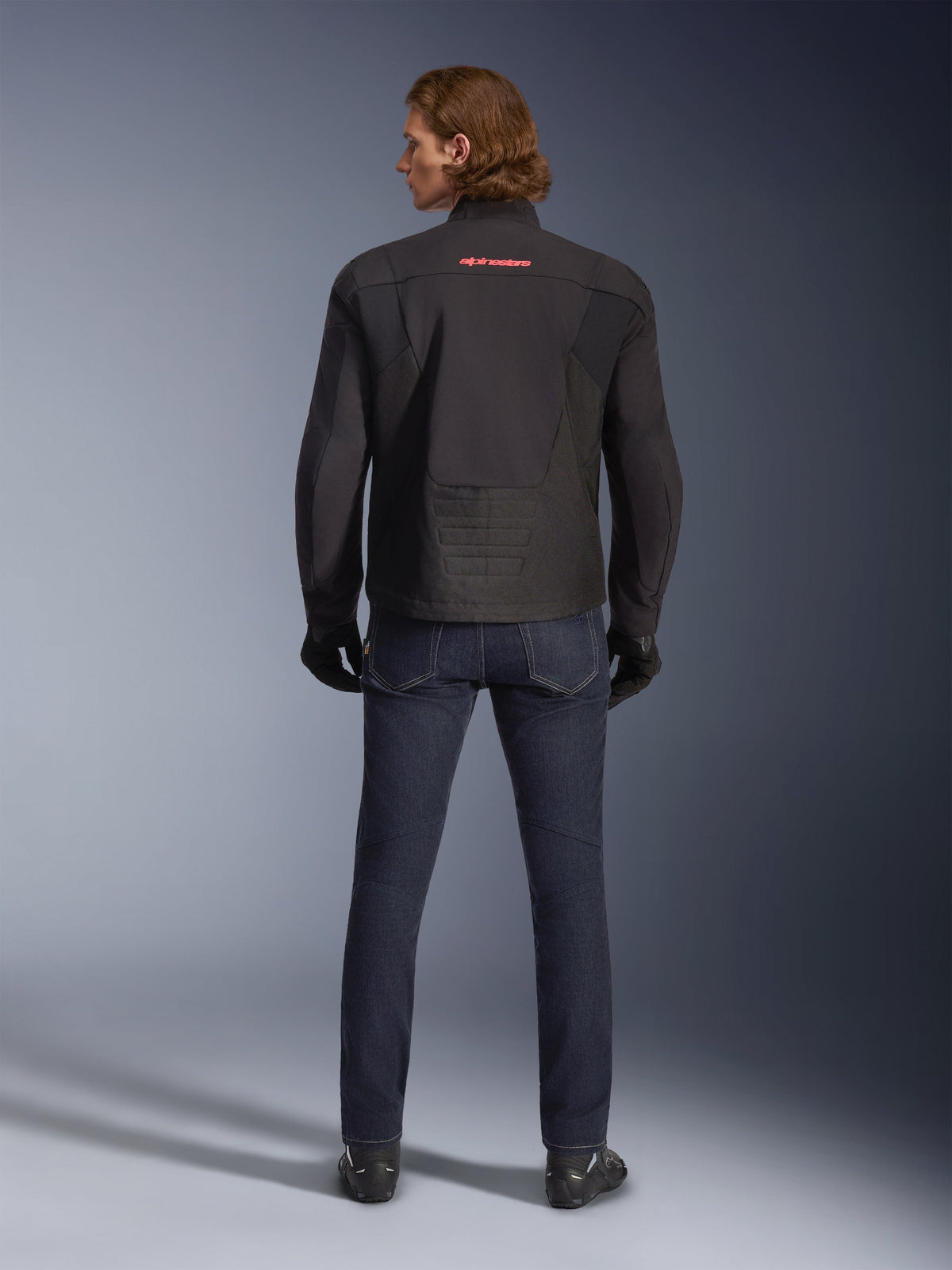 Chaqueta impermeable Alpinestars SMX, negra, vista trasera usada por un modelo, con un logotipo rojo en la parte superior de la espalda, dobladillo trasero alargado para mayor cobertura al conducir, combinada con vaqueros de moto azul oscuro y guantes negros.