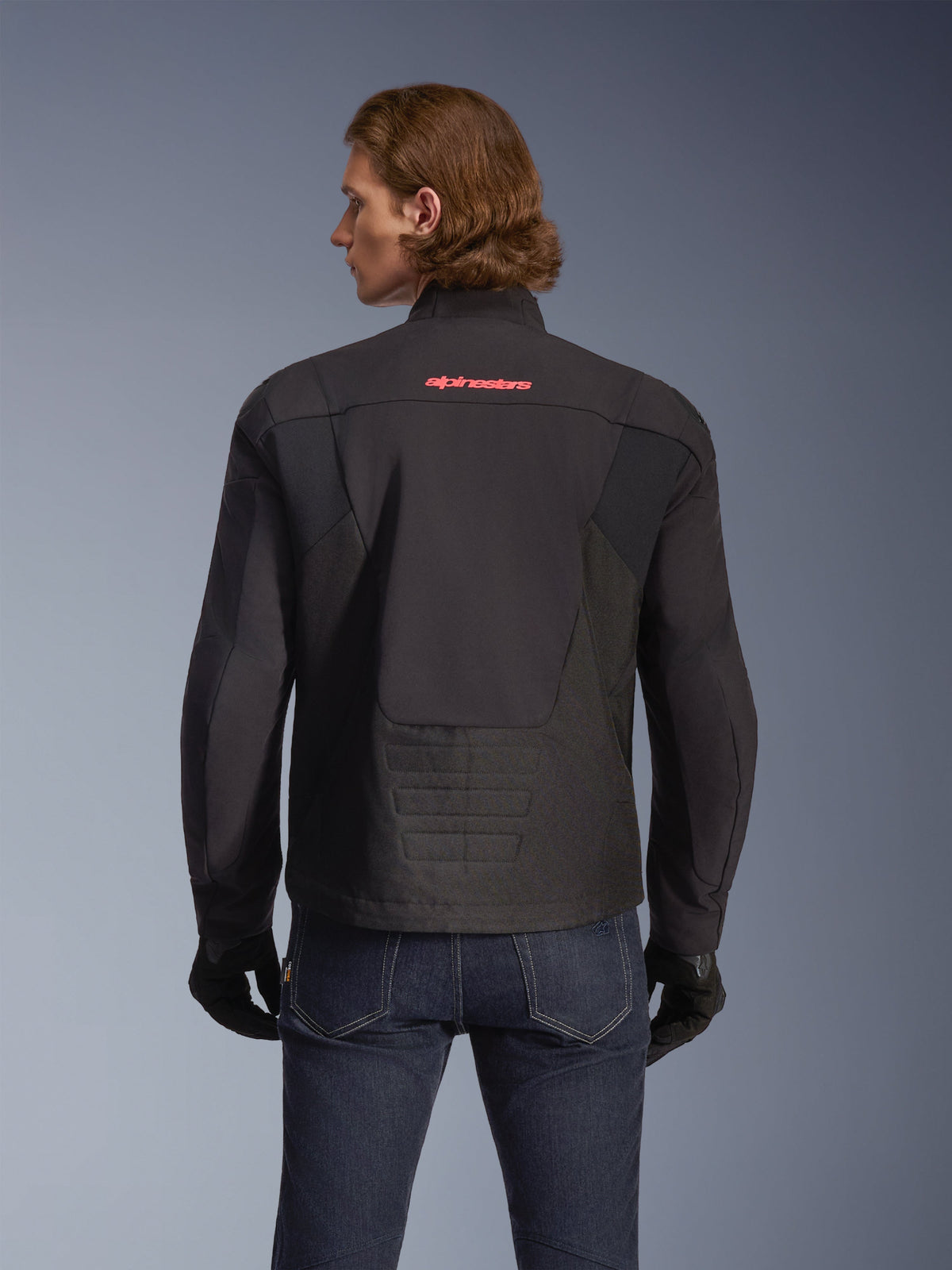 Chaqueta impermeable Alpinestars SMX, negra con logotipo en rojo brillante, vista trasera mostrada en un modelo, combinada con vaqueros azul oscuro y guantes negros, chaqueta de moto textil para conducción en carretera y deportiva.