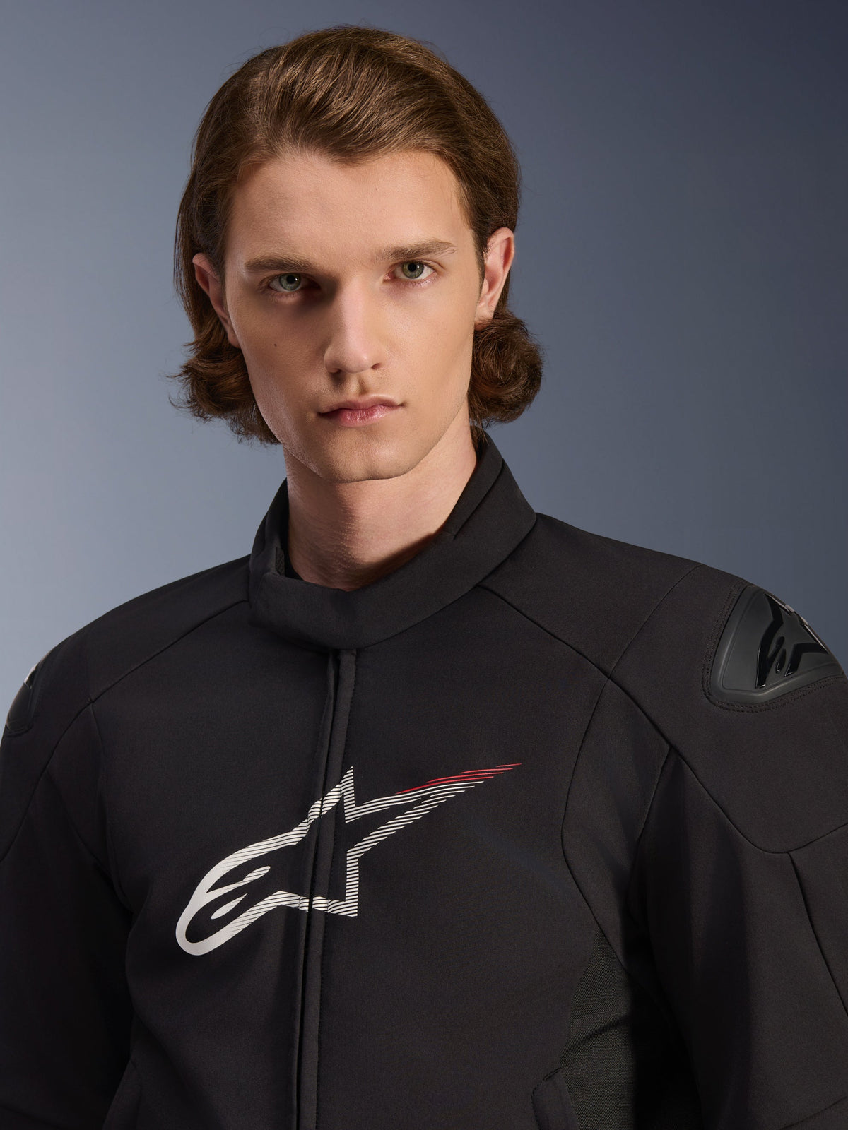 Modelo con la Chaqueta Impermeable Alpinestars SMX, color negro con detalles en blanco y rojo brillante, que presenta un gran logotipo de estrella blanca con una franja roja en el pecho, protectores de hombros DFS Lite y una construcción textil para el rendimiento en carretera.