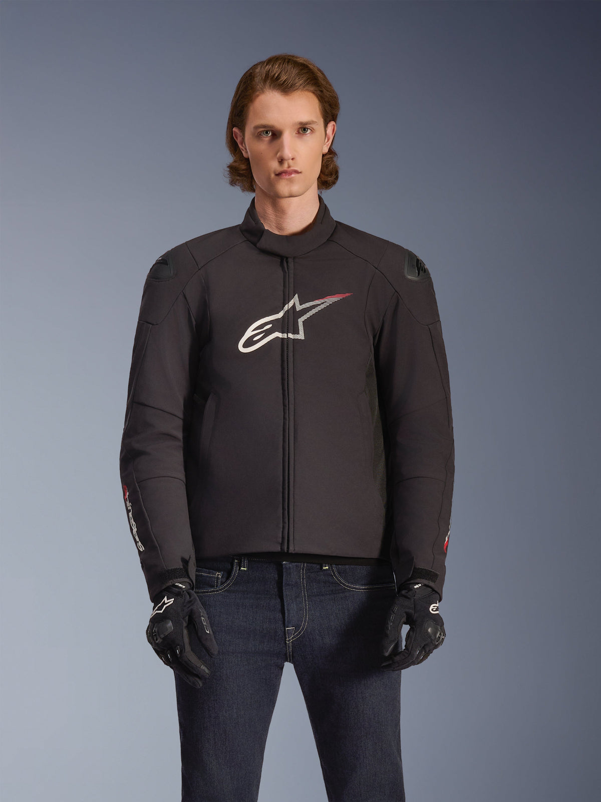 Chaqueta impermeable Alpinestars SMX, chaqueta de moto textil negra con logotipo de estrella en blanco y rojo en el pecho, lucida por un modelo con guantes negros y vaqueros azules, con deslizadores externos en los hombros y un ajuste deportivo.