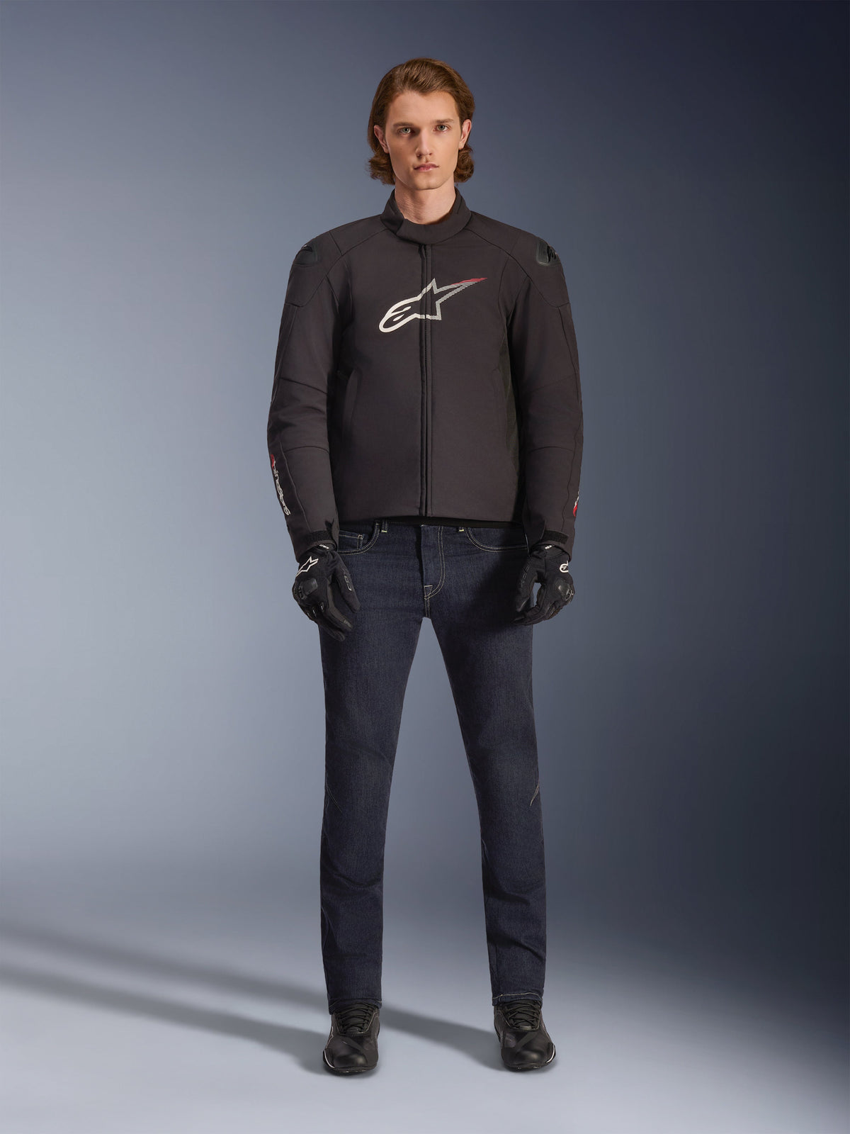 Modelo con la chaqueta impermeable Alpinestars SMX en color negro con logotipo blanco y rojo, combinada con pantalones de montar de denim oscuro, guantes negros y botas de conducción, conjunto deportivo de equipamiento de motociclismo para todo clima.