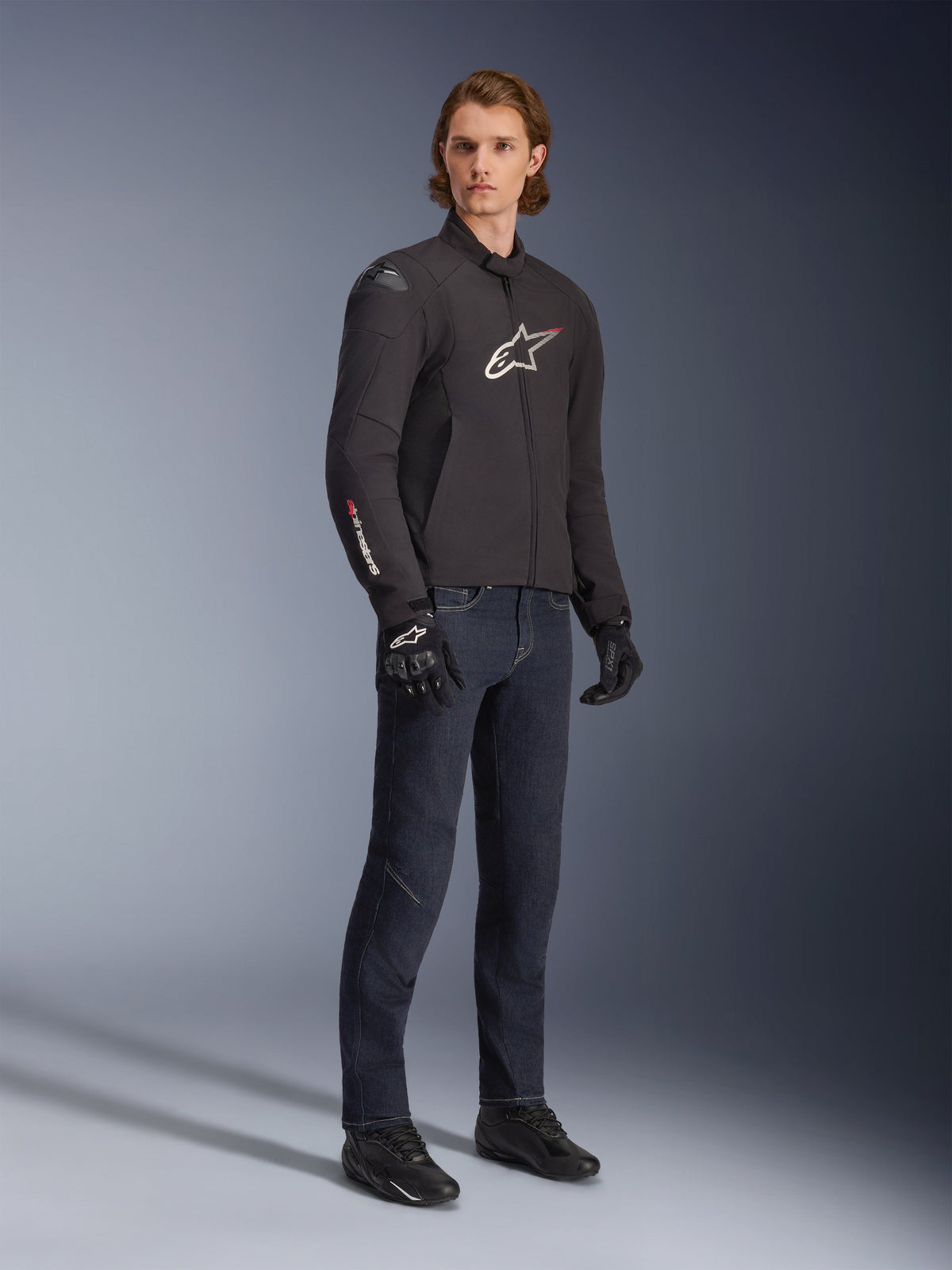 Modelo masculino con chaqueta Alpinestars SMX Waterproof Jacket, negra con detalles de logotipos en blanco y rojo, combinada con pantalones denim azul oscuro y guantes de conducción negros, chaqueta de moto textil de corte deportivo.