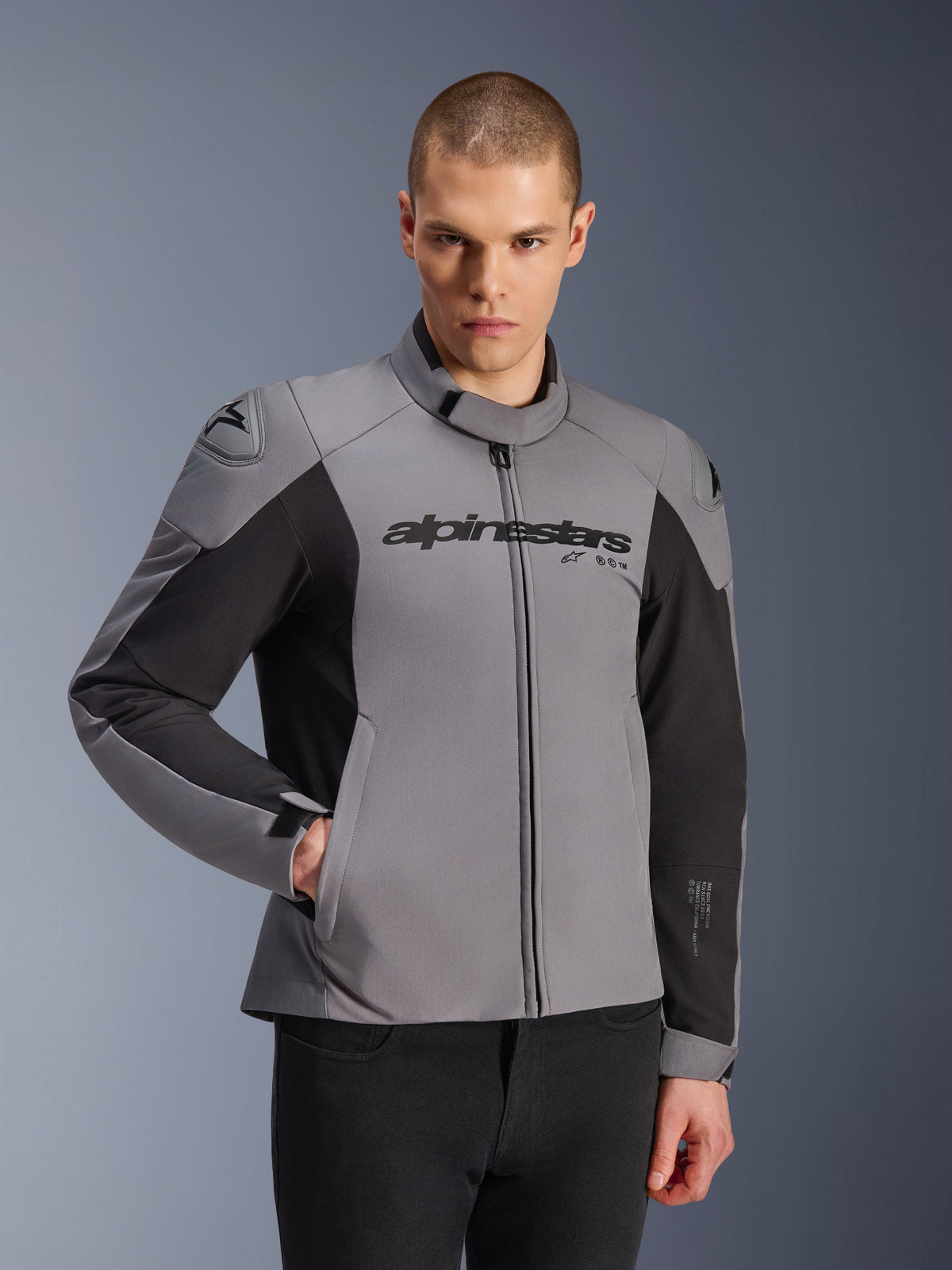 Chaqueta impermeable Alpinestars SMX, gris oscuro y negro, usada por un modelo masculino, con el logotipo 'alpinestars' en el pecho, protección de hombros DFS Lite, diseño deportivo elegante con materiales textiles y softshell para conducción en carretera.