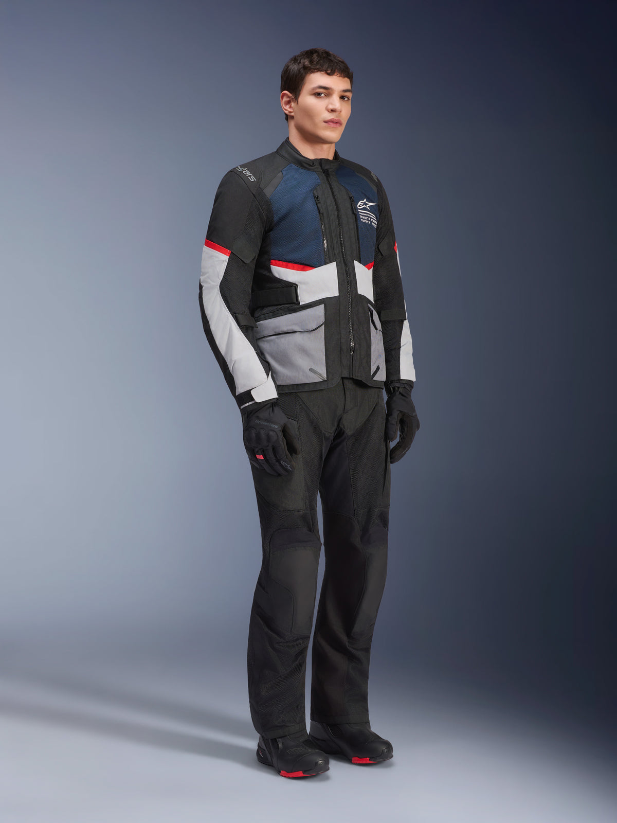 Modelo masculino con la chaqueta Alpinestars Andes Air Drystar® en azul profundo, negro y gris hielo, combinada con pantalones, guantes y botas de moto negros, con amplios paneles de malla para ventilación, bolsillos gris claro y detalles en rojo, diseñada para touring de aventura.