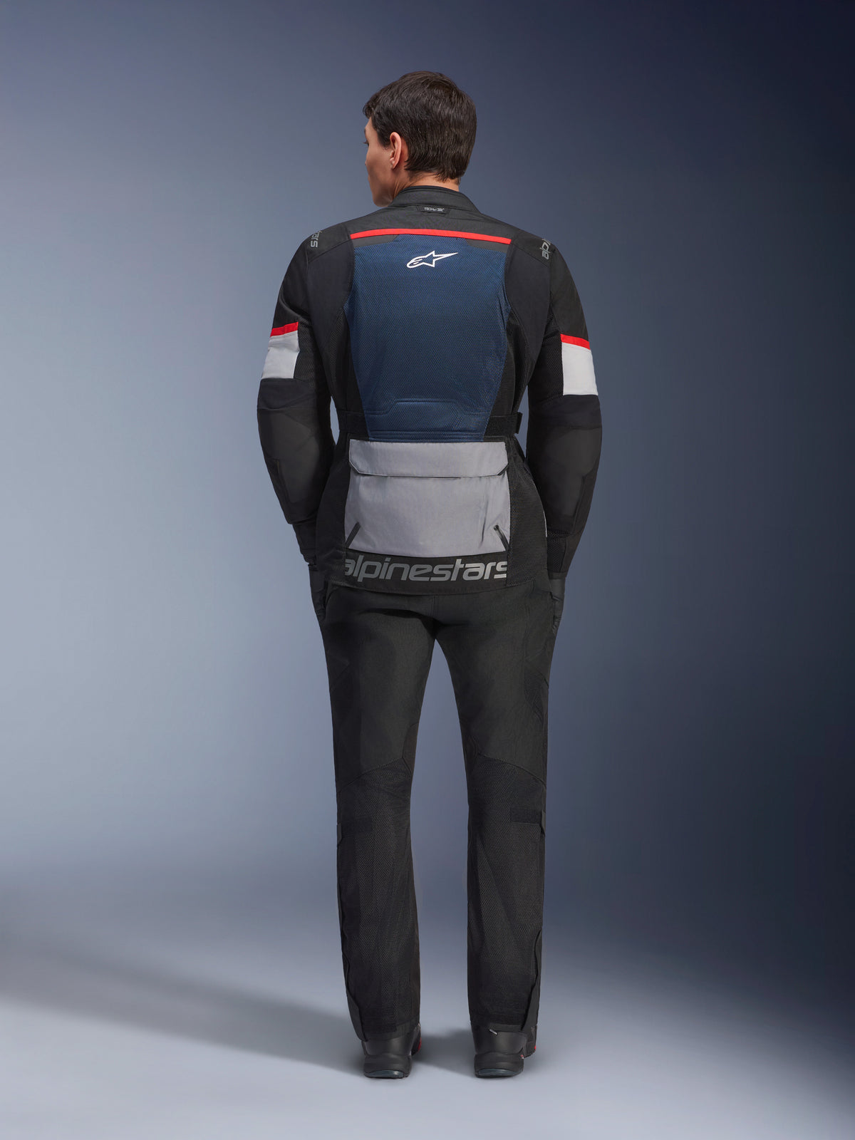 Chaqueta Alpinestars Andes Air Drystar, Deep Blue Black Ice Gray, vista posterior en modelo, con panel de malla azul en la espalda para ventilación, bolsillo inferior gris de gran tamaño, detalles en rojo y blanco en las mangas y logotipo 'alpinestars', combinada con pantalones de moto negros para un conjunto de touring completo