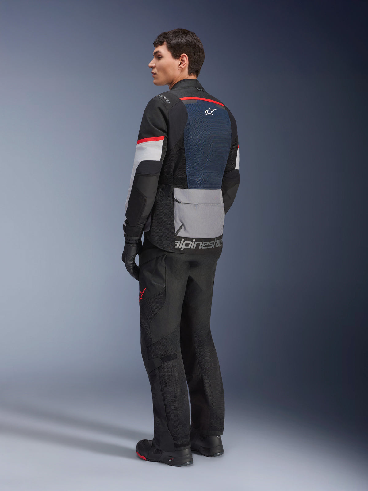 Modelo masculino en vista lateral trasera vistiendo la chaqueta Alpinestars Andes Air Drystar® en color Deep Blue Black Ice Gray, combinada con pantalones y guantes de moto negros, con un panel de malla azul en la espalda, bolsillo utilitario gris en la parte baja de la espalda y detalles rojos en las mangas, equipo profesional de adventure touring.