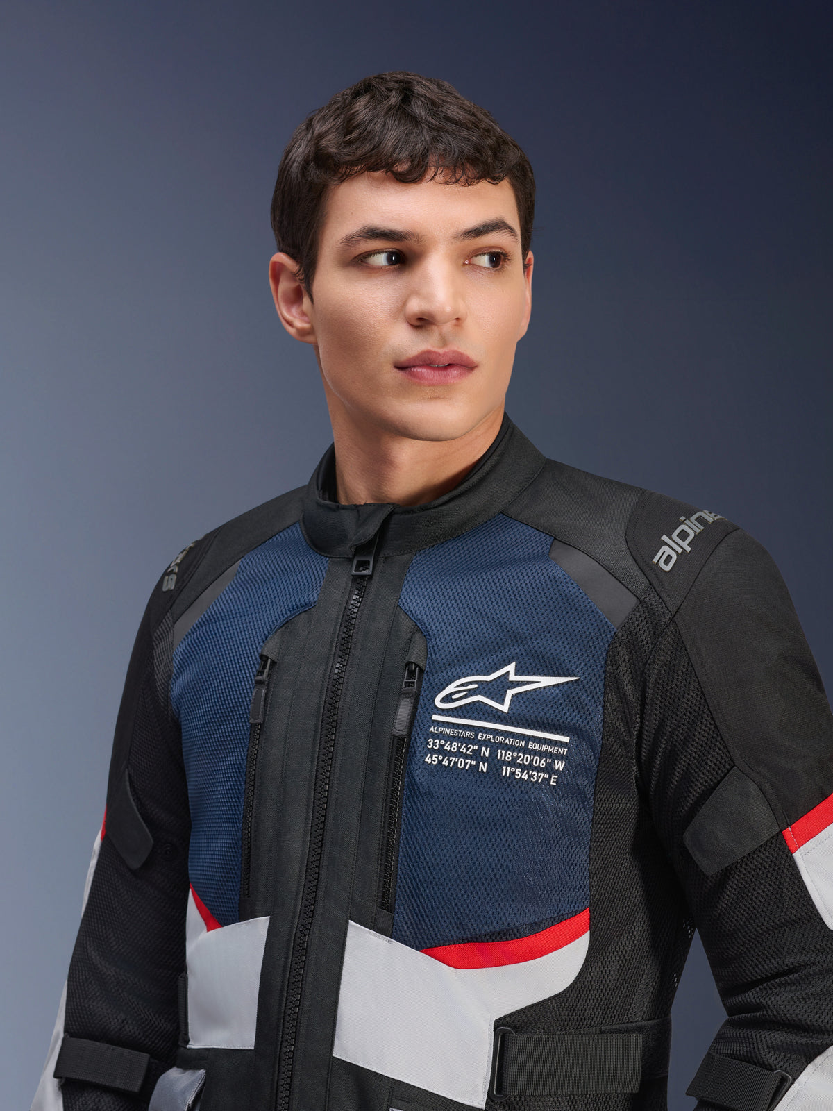 Chaqueta Alpinestars Andes Air Drystar®, chaqueta de moto, en azul oscuro, negro y gris hielo con detalles rojos, usada por un modelo masculino, con paneles de malla ventilada en el pecho, logotipo de la estrella blanca con coordenadas de exploración y diseño de aventura listo para la carretera