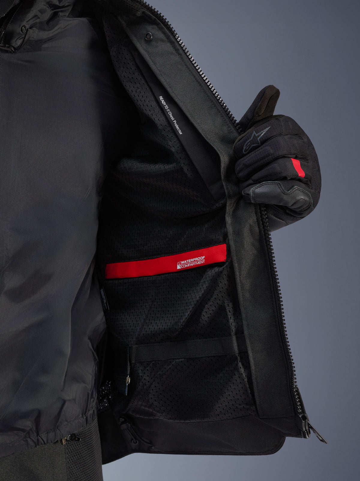 Vista detallada del interior de la chaqueta Alpinestars Andes Air Drystar®, destacando el forro de malla negra transpirable, el compartimento impermeable rojo y el bolsillo para el protector de pecho, mostrada siendo sostenida abierta por un guante de moto negro.