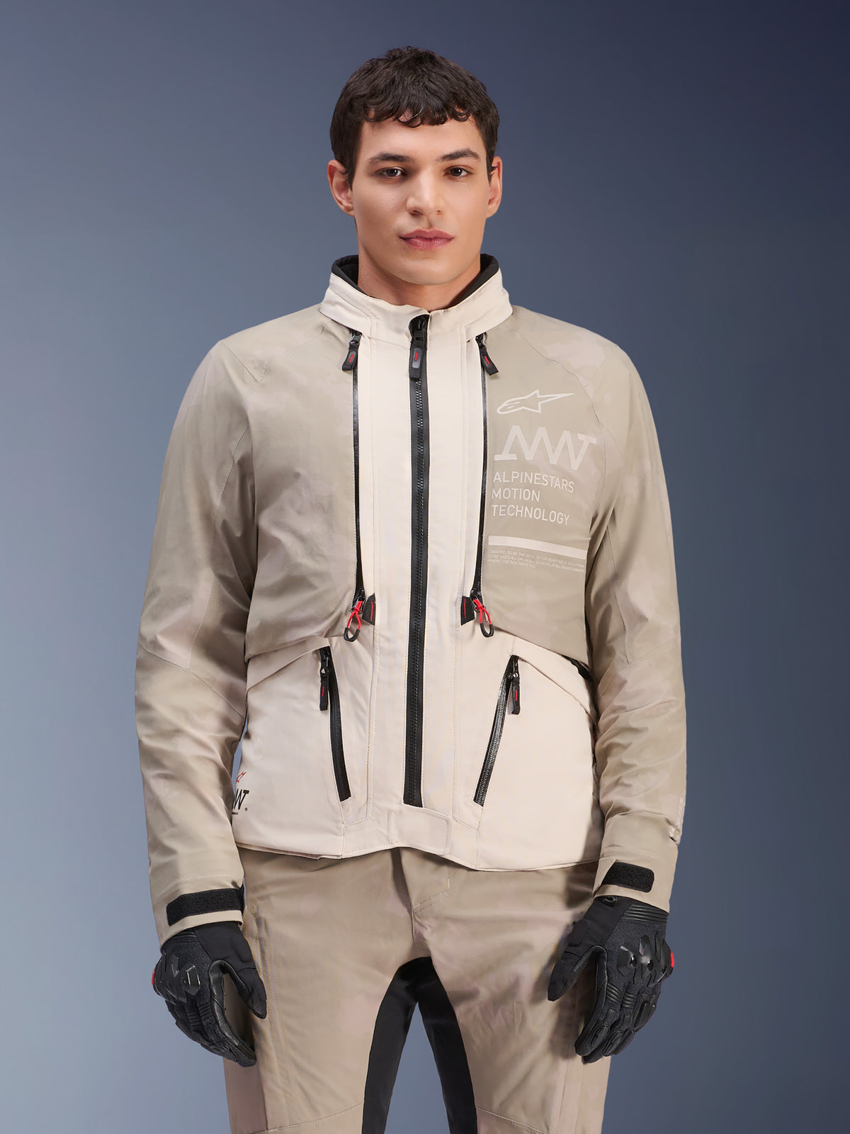 Chaqueta Alpinestars AMT-10LAB Drystar® XF, chaqueta de moto adventure, White Sand Aluminum Camo, diseño modular con cremalleras negras y acentos rojos, usada con pantalones a juego y guantes negros, con logotipo Alpinestars Motion Technology en el pecho, equipo de conducción profesional para todo tipo de clima