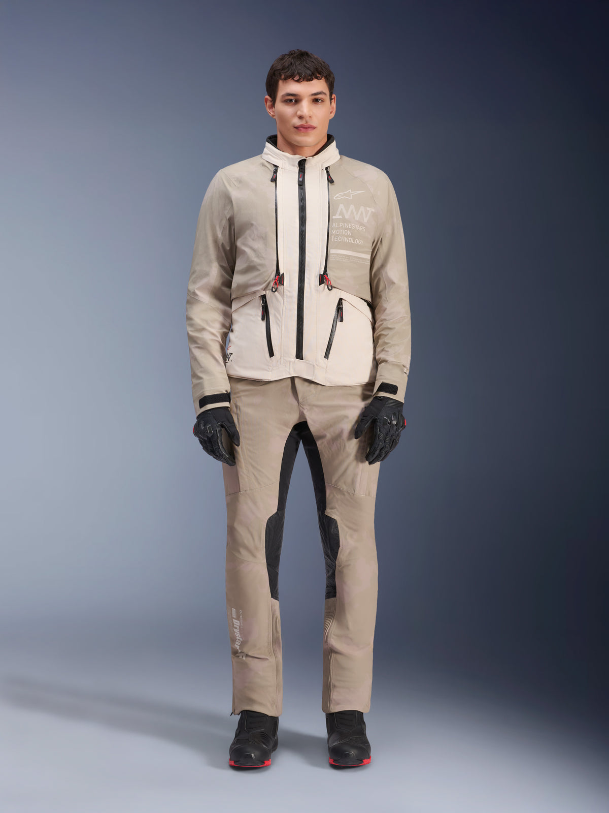 Chaqueta Alpinestars AMT-10LAB Drystar® XF en White Sand Aluminum Camo, usada por un modelo con pantalones de aventura a juego, guantes negros y botas negras, equipamiento técnico modular de moto para touring multitemporada.