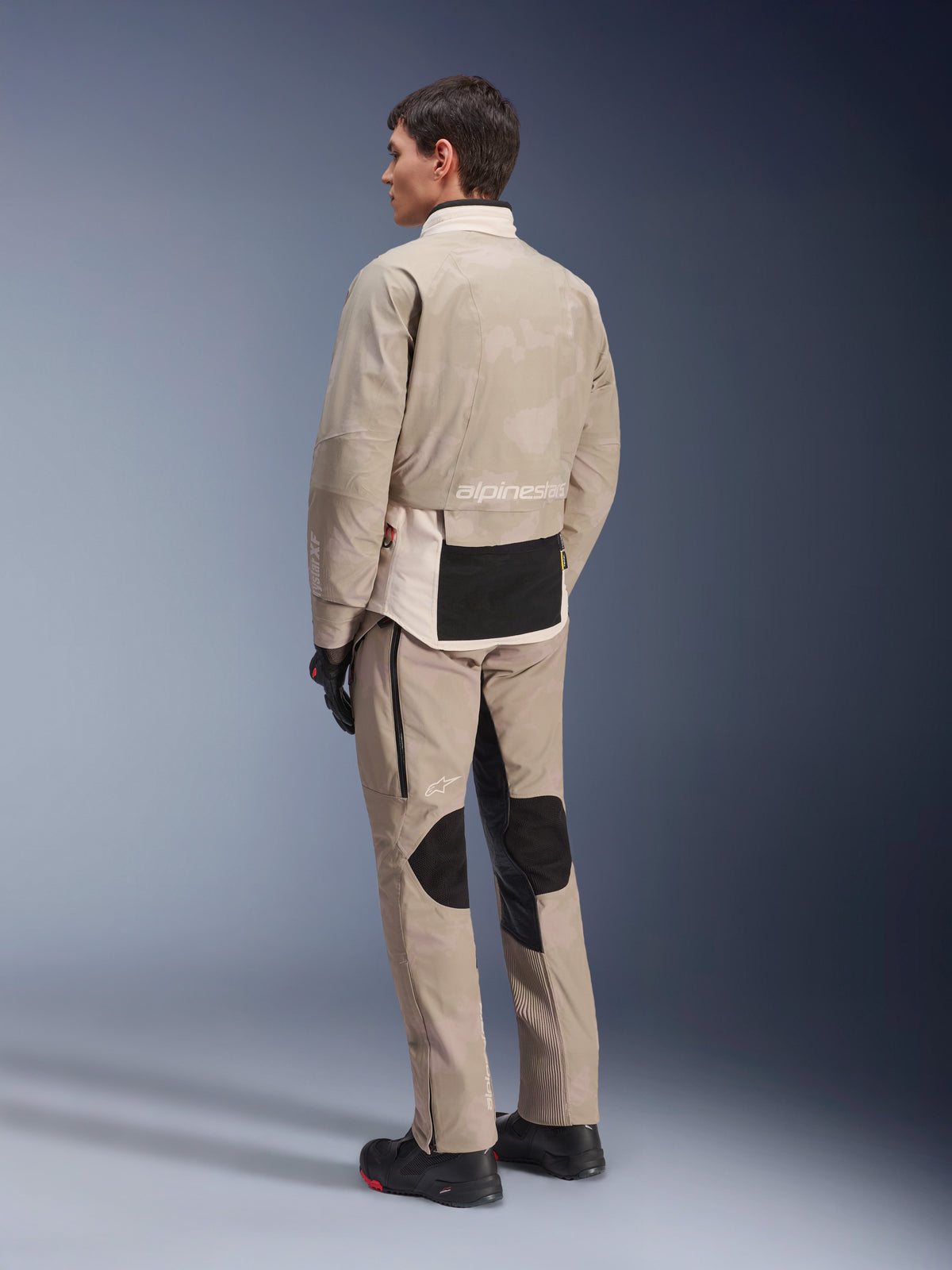 Modelo desde atrás vistiendo la chaqueta AMT-10LAB Drystar® XF y pantalones a juego, White Sand Aluminum Camo, equipamiento de aventura para motociclismo, con paneles utilitarios negros y marca Alpinestars, combinado con botas y guantes negros de motorista, conjunto coordinado para touring en cualquier condición climática