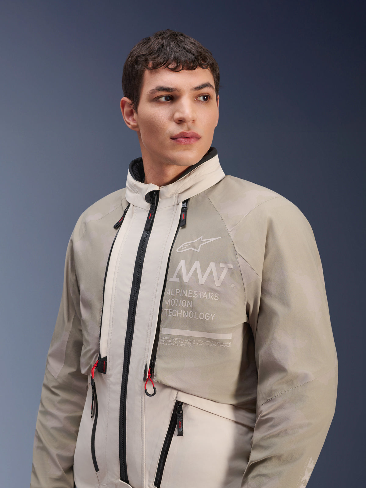 Chaqueta Alpinestars AMT-10LAB Drystar® XF, White Sand Aluminum Camo, chaqueta modular de aventura para motociclista, con la marca "Alpinestars Motion Technology" en el pecho, usada por un modelo masculino.